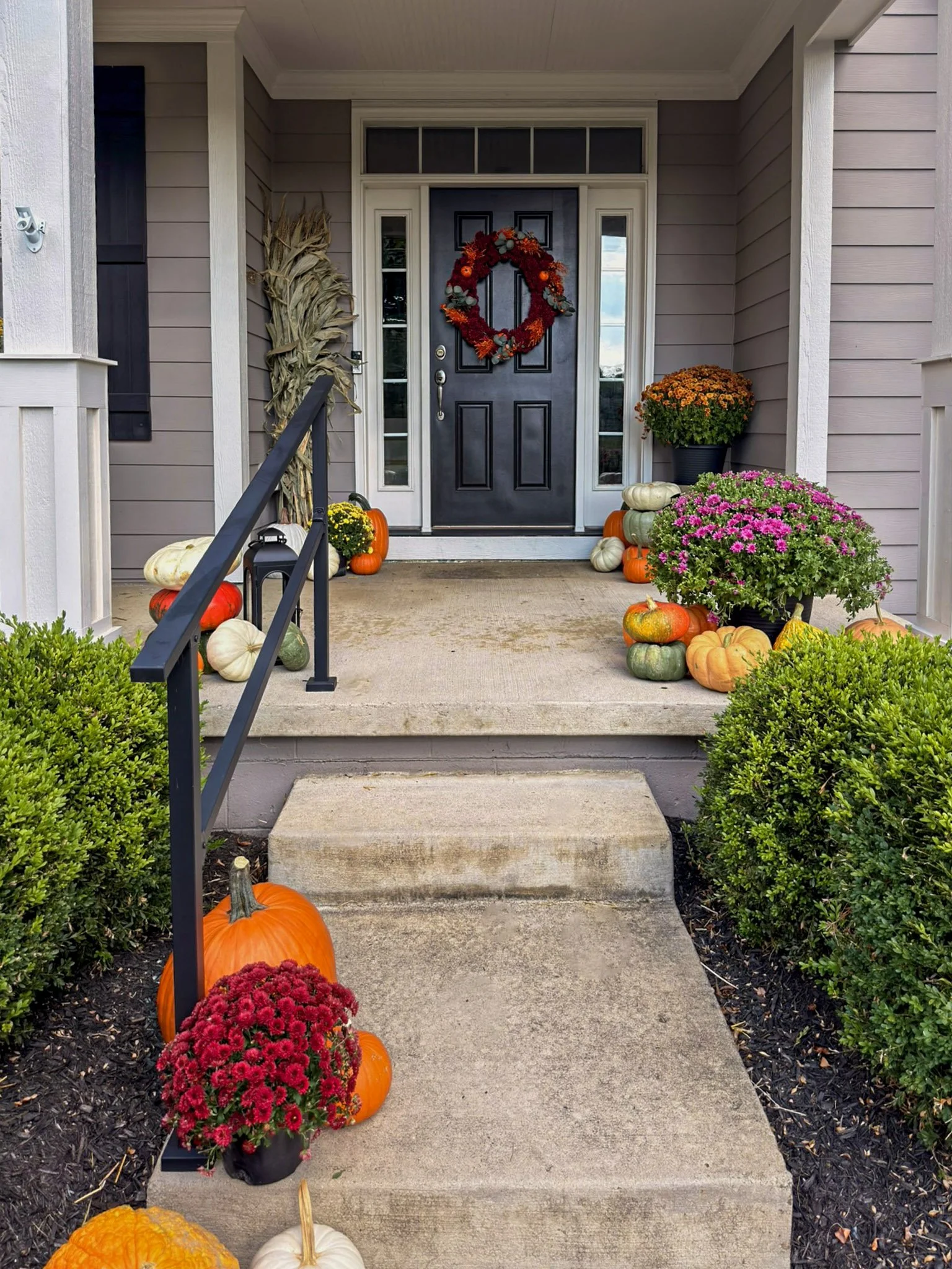 Dark front door with a red wreath, black railing, corn stalks, mums and pumpkins flanking the walkway