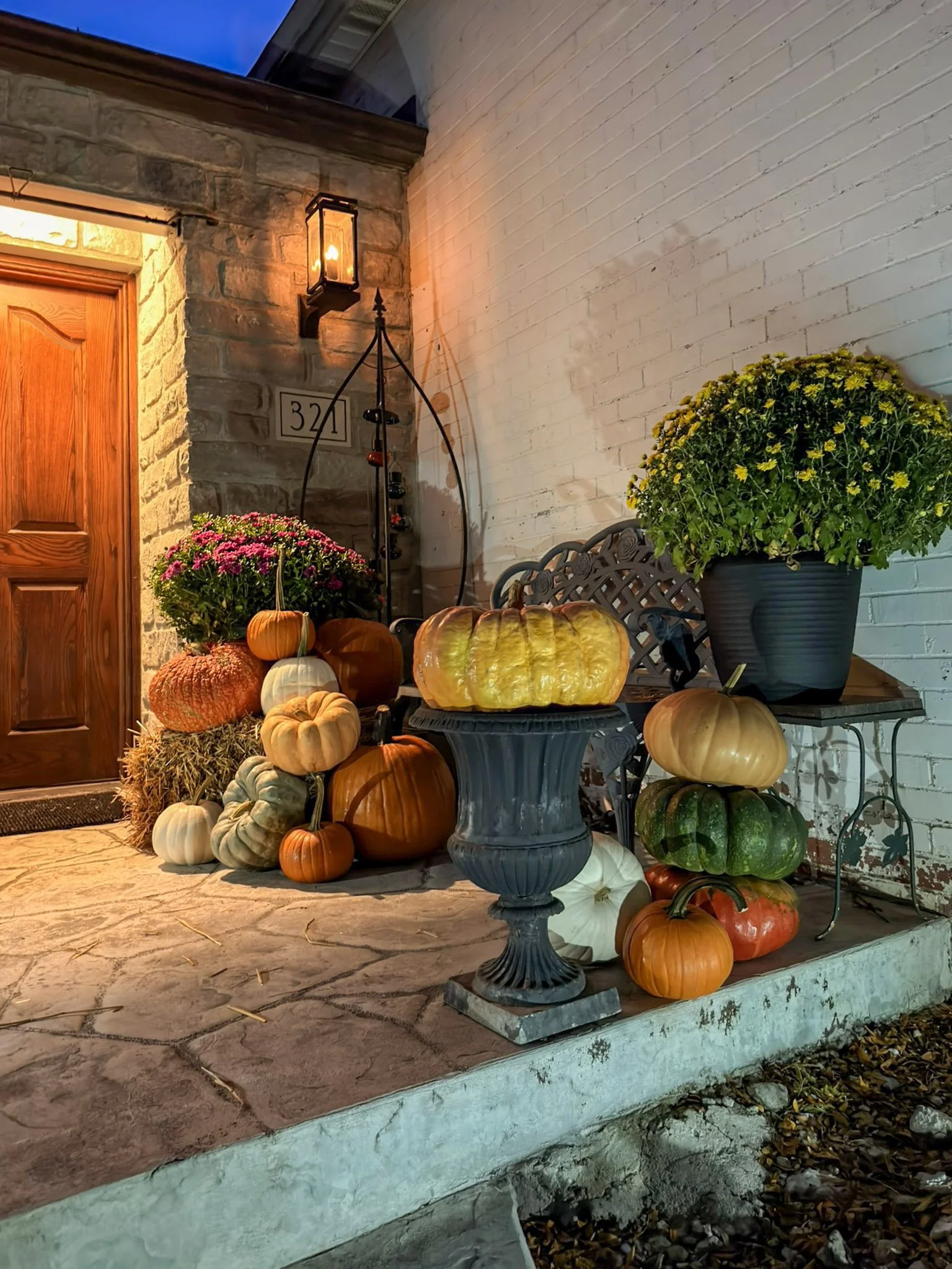 Hay bale, urn of mums and mixed orange and heirloom pumpkins styled beside a stone entry at dusk