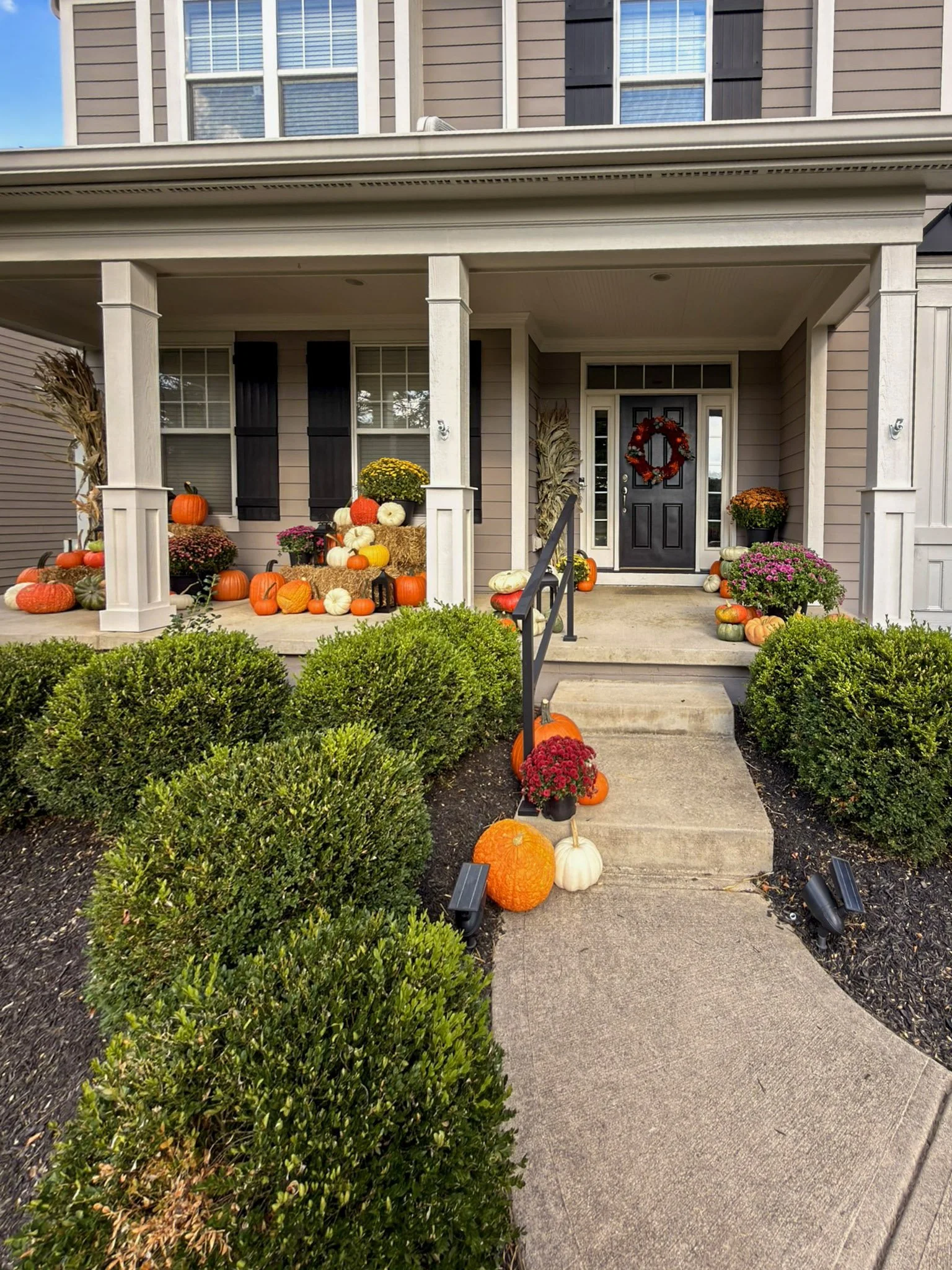 Two story home with white porch columns, a red door wreath, corn stalks and pumpkins along the porch