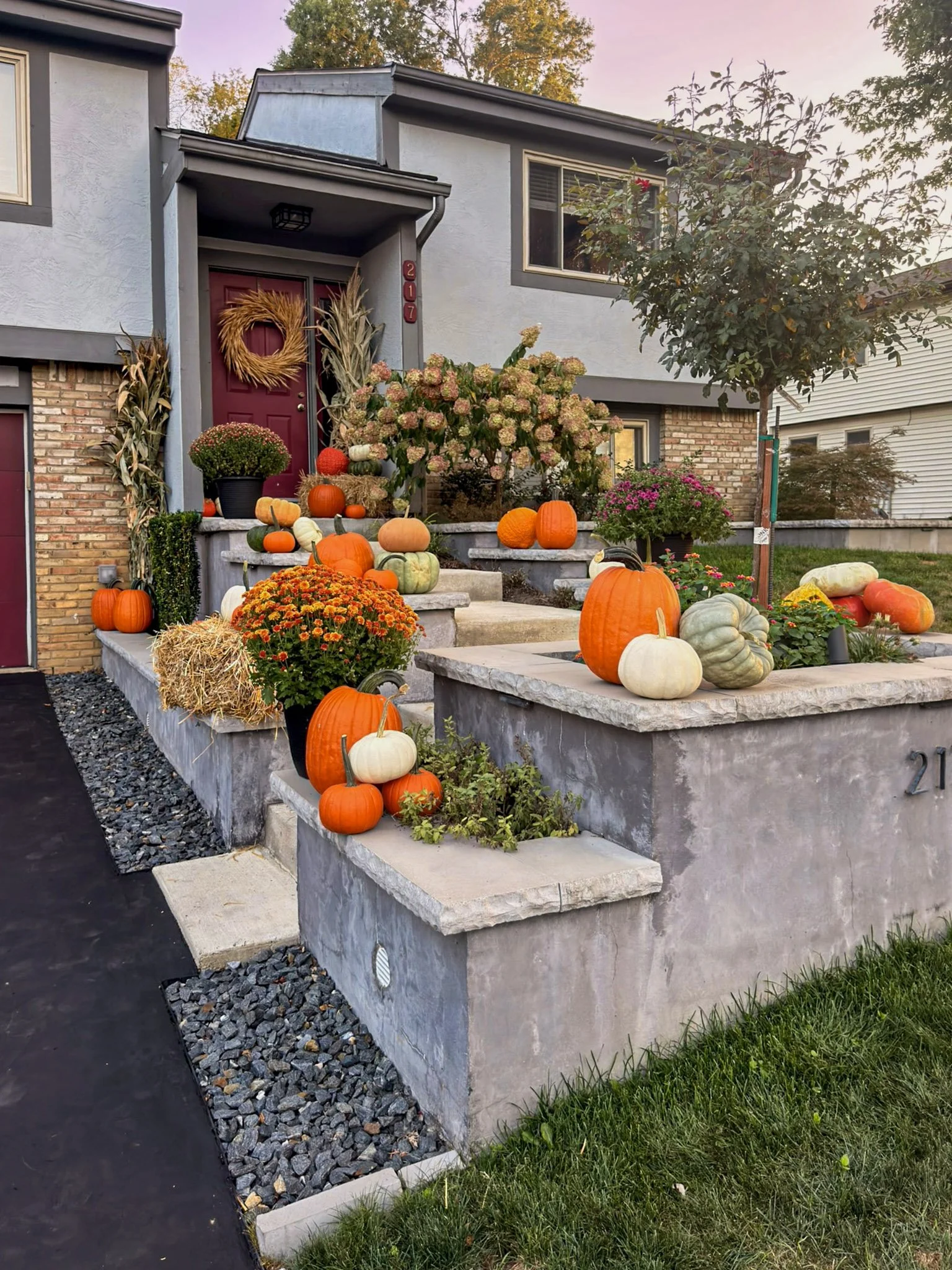 Red front door with a fall wreath, corn stalks, mums and pumpkins on a raised concrete porch wall