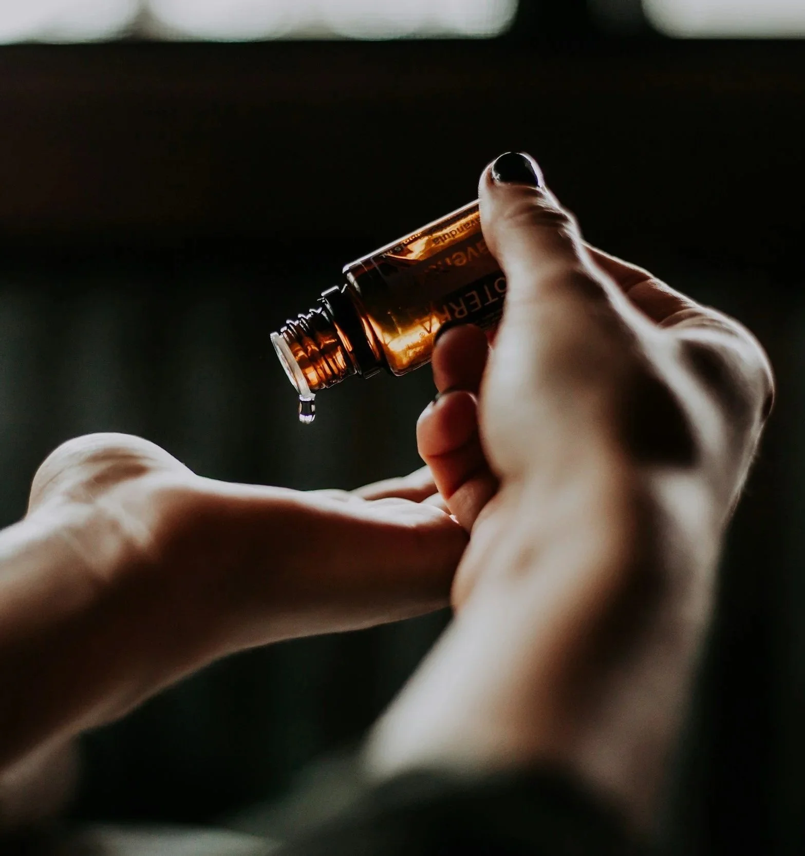 A person holding a small amber glass bottle with a black and orange label, pouring a liquid into their hand in a dimly lit setting.