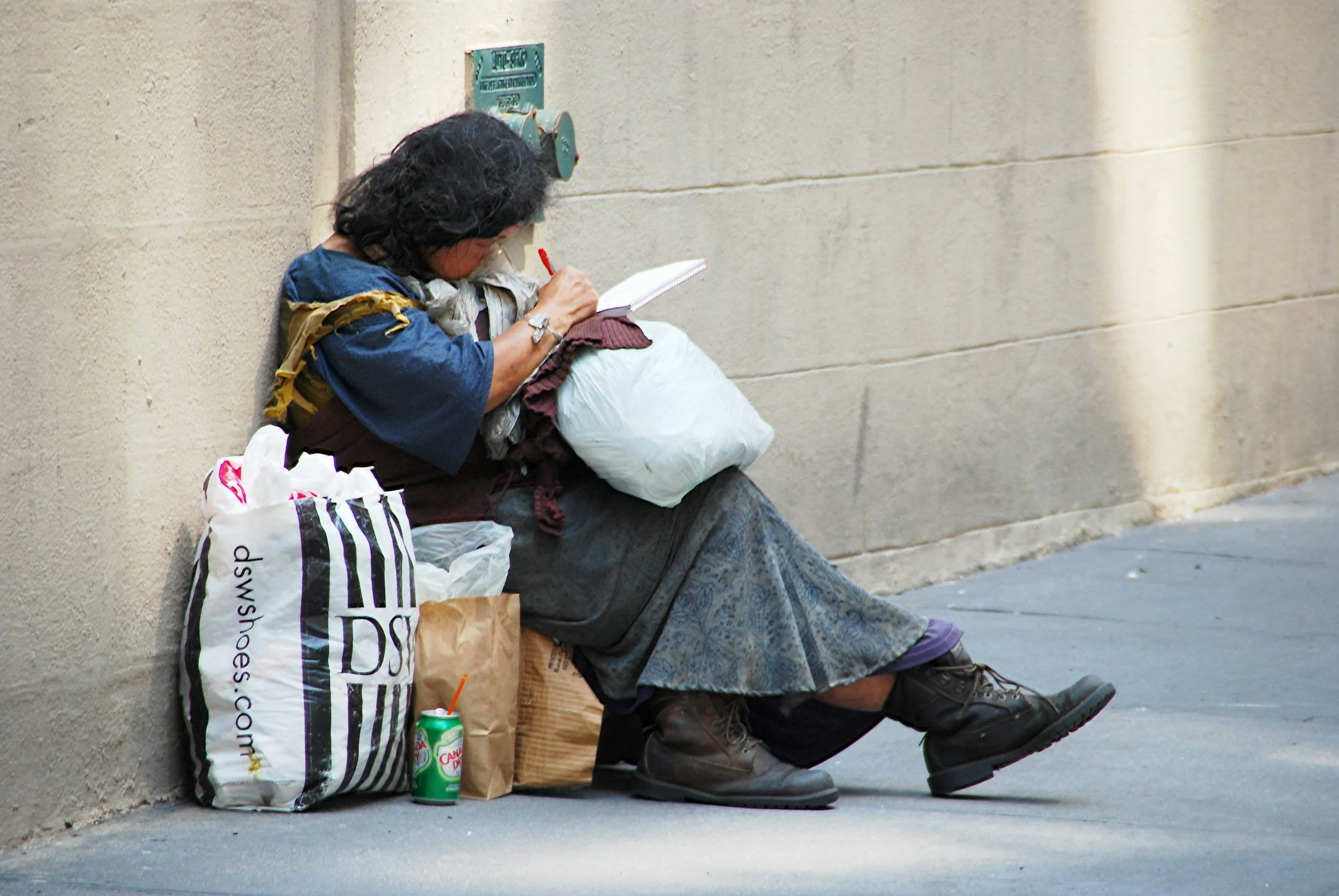 A person with dark, curly hair sitting on a sidewalk against a beige wall, reading a book. They are wearing a blue shirt, a gray scarf, and gray pants. There are shopping bags and a soda can on the ground next to them.