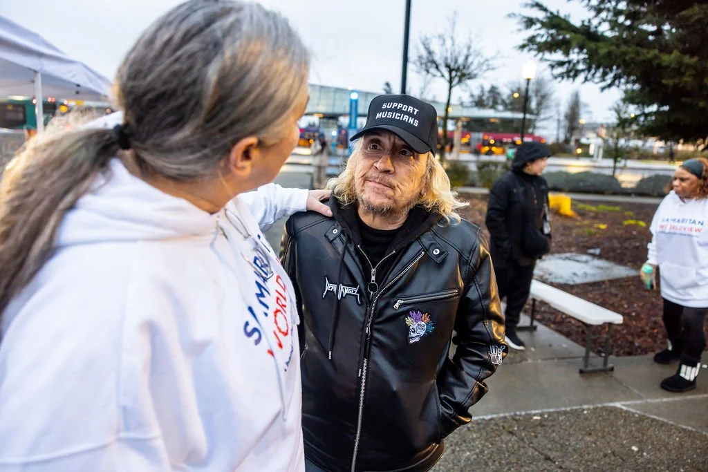 Two people having a conversation outdoors on a rainy day, with others in the background. One person has gray hair tied in a ponytail and is wearing a white jacket; the other has long blond hair, is wearing a black leather jacket, and a black cap that says 'Support Musicians.'