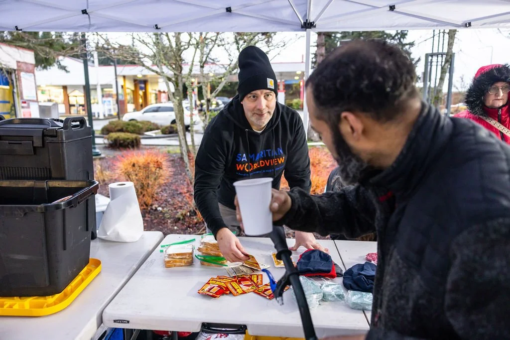 A man in a black beanie and sweatshirt distributes hot dogs at a table under a white tent, with a customer holding a cup in front of him.
