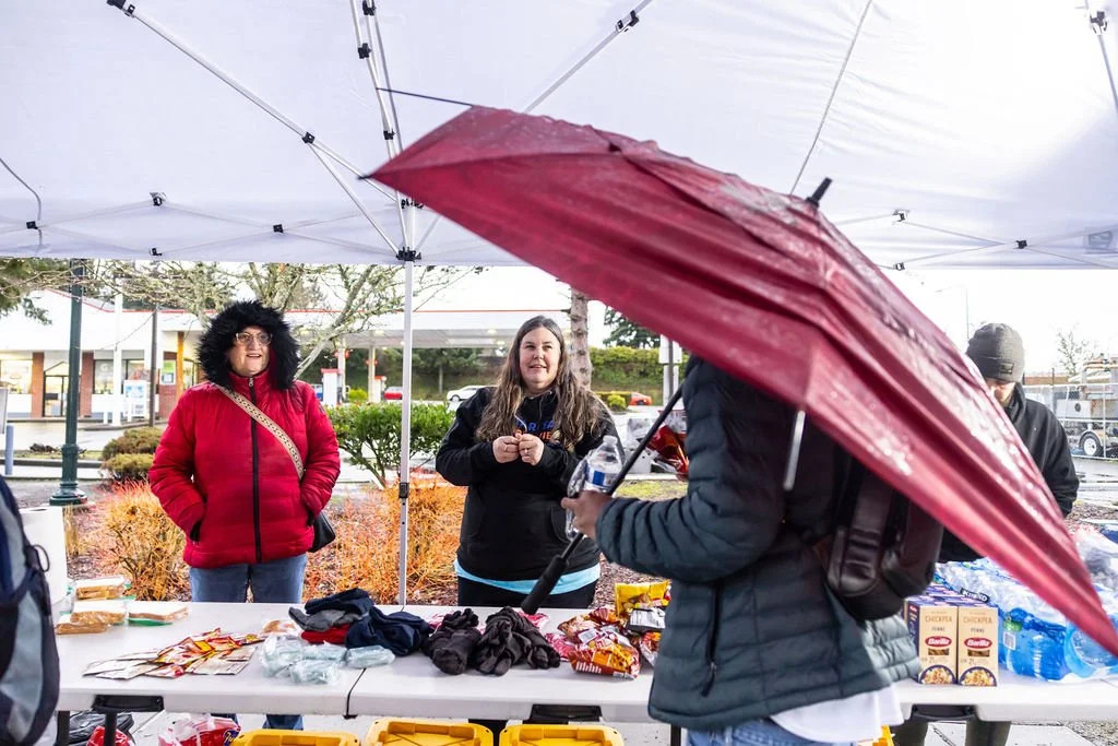 People are gathered under a white canopy at an outdoor event in the rain. One woman is smiling and talking, another woman is standing nearby, and a man is holding a red umbrella and a water bottle. There is a table with snacks and other items in front of them.