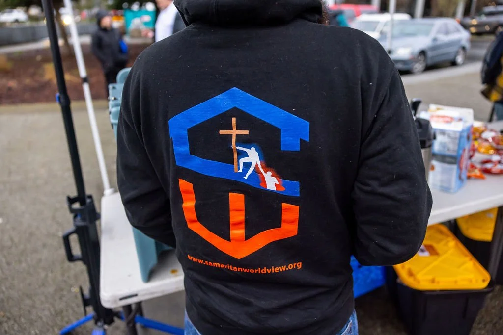 Person wearing a black hoodie with a logo and the website samaritans.org on the back, standing near a table with food at an outdoor event.