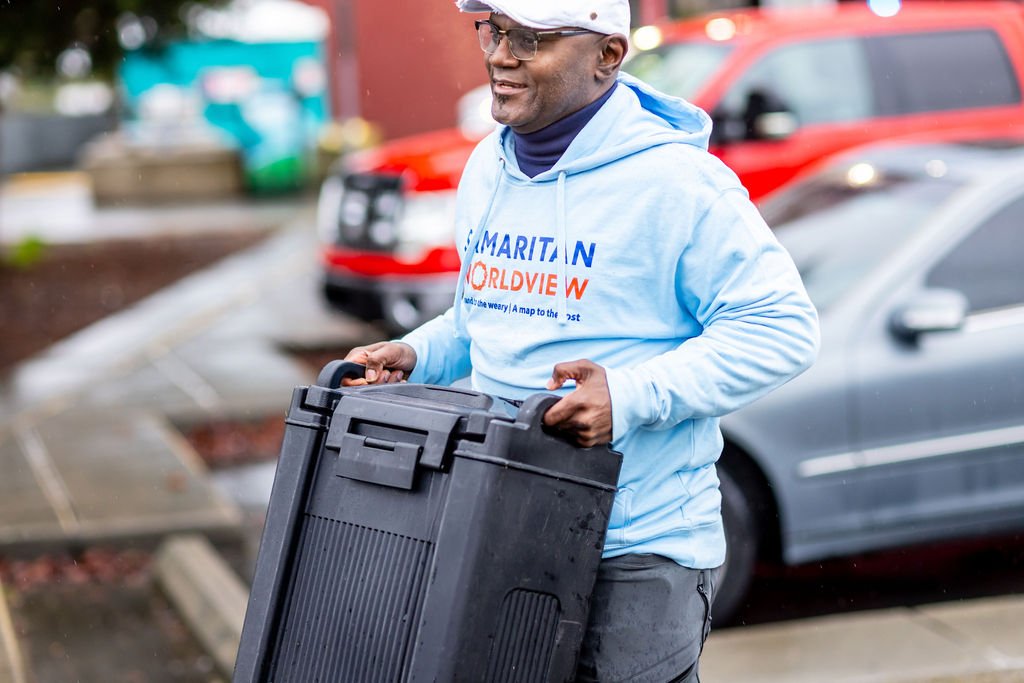A man wearing glasses, a white cap, and a light blue hoodie that reads 'Maritime' is holding a large black case while walking outdoors near parked cars.