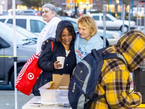 Two women smiling while one holds a cup, at a table with food containers, with a young person in a yellow plaid hoodie and backpack in the foreground, outside in a parking lot.