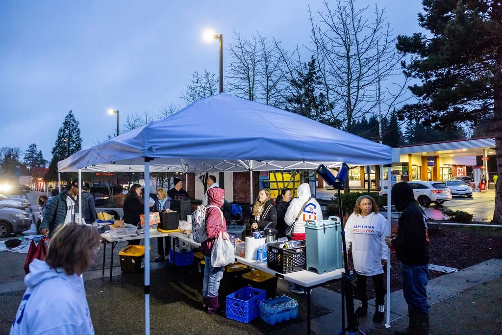 A group of people gathered under a white canopy tent in a parking lot during the evening. There are tables with supplies, and some people are talking or working. In the background, there are parked cars, a gas station, and leafless trees.