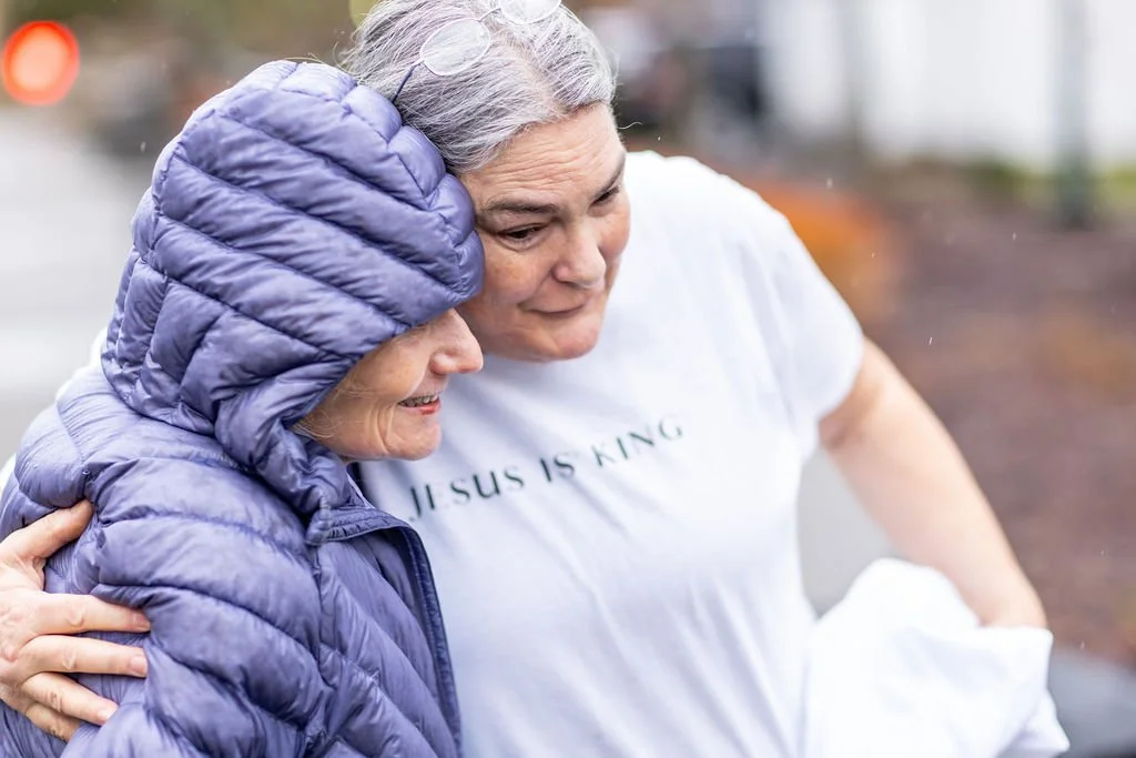 Two women hugging outdoors, one wearing a purple puffer jacket with the hood up, and the other wearing a white T-shirt with text that reads 'JESUS IS KING'.