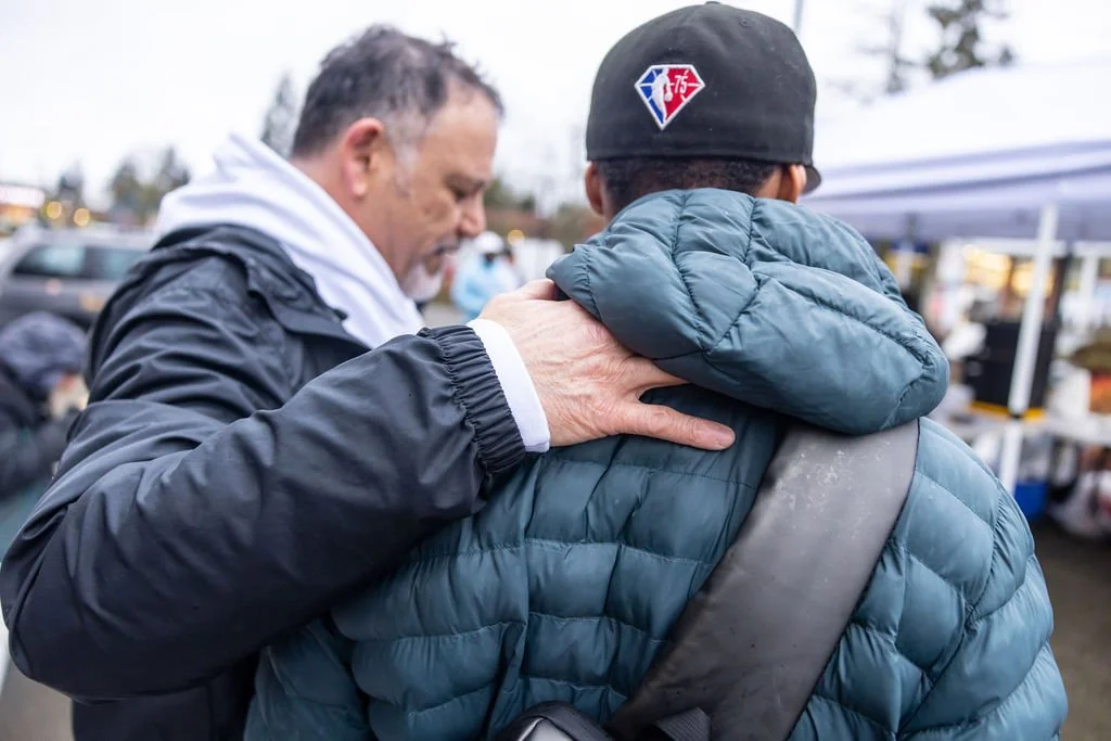 Two men greet each other at an outdoor market, with one man placing his hand on the other's shoulder. One man wears a black cap and blue puffer jacket, the other wears a dark jacket with a white hoodie.