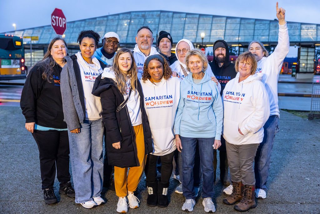 Group of diverse people wearing Samaritan Worldview hoodies standing outdoors in rainy weather near buses and a stop sign, smiling and posing for a photo.