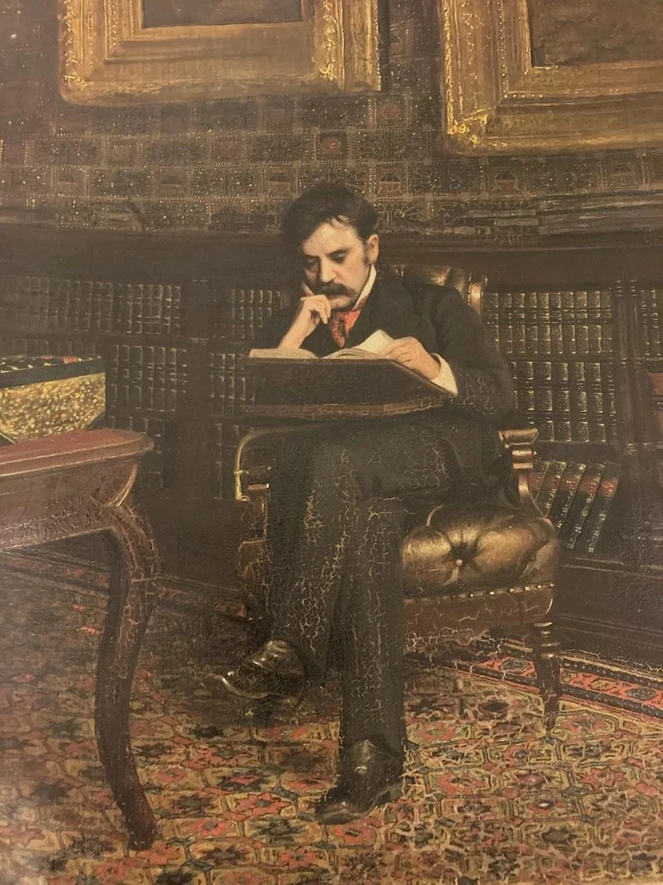 A man with dark hair and a mustache, dressed in vintage clothing, sits in an ornate, vintage armchair in a richly decorated room, reading a book.