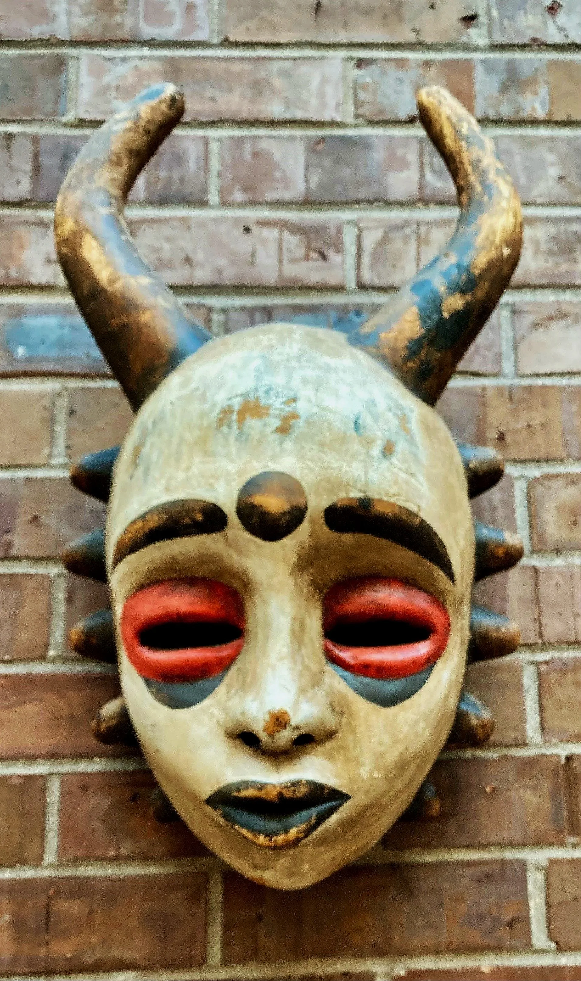 Decorative tribal mask with antler-like horns mounted on a brick wall.