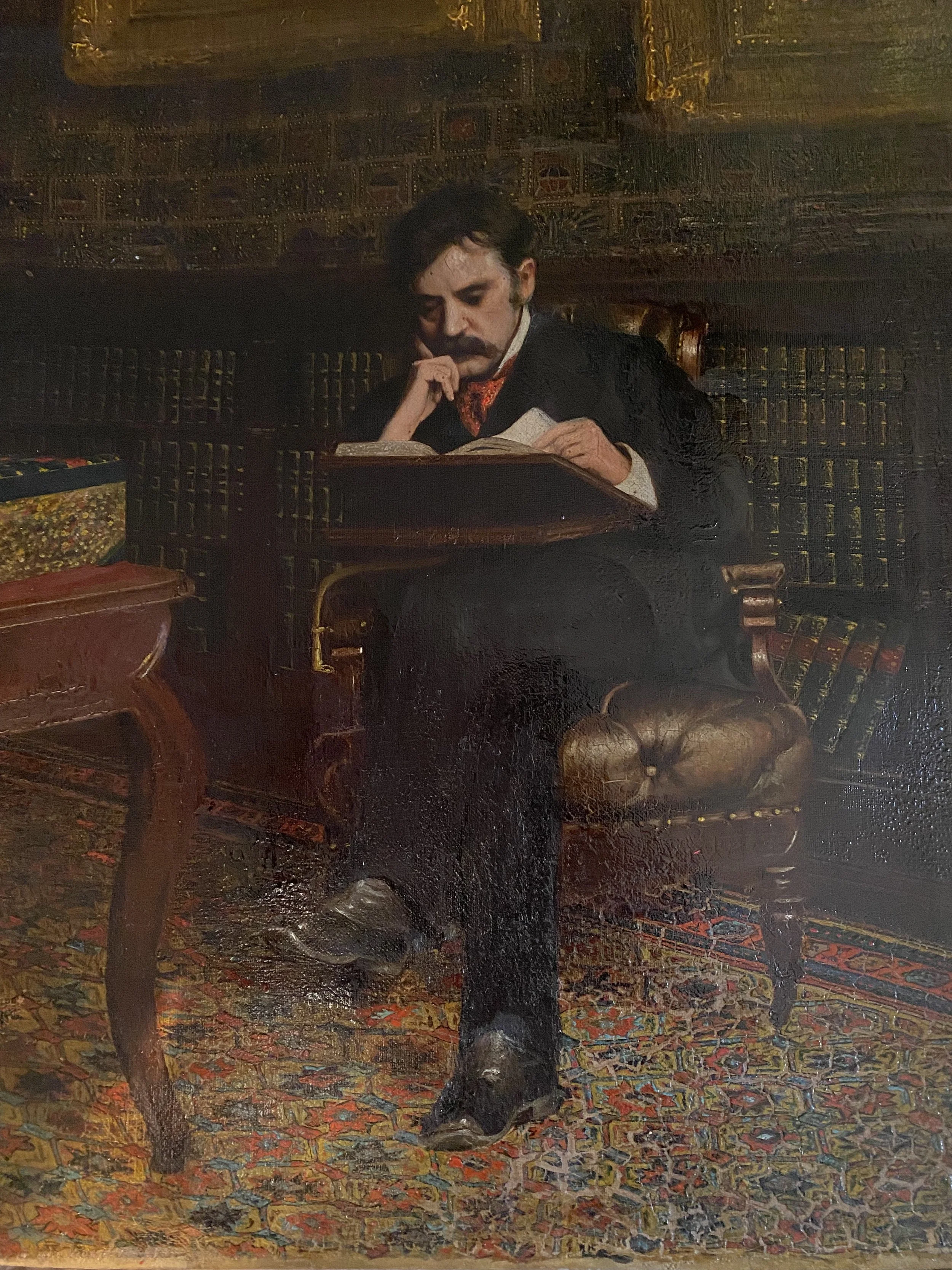 Painting of a man with dark hair and mustache sitting in a leather armchair, reading a book, in a room with dark wooden bookshelves filled with books.