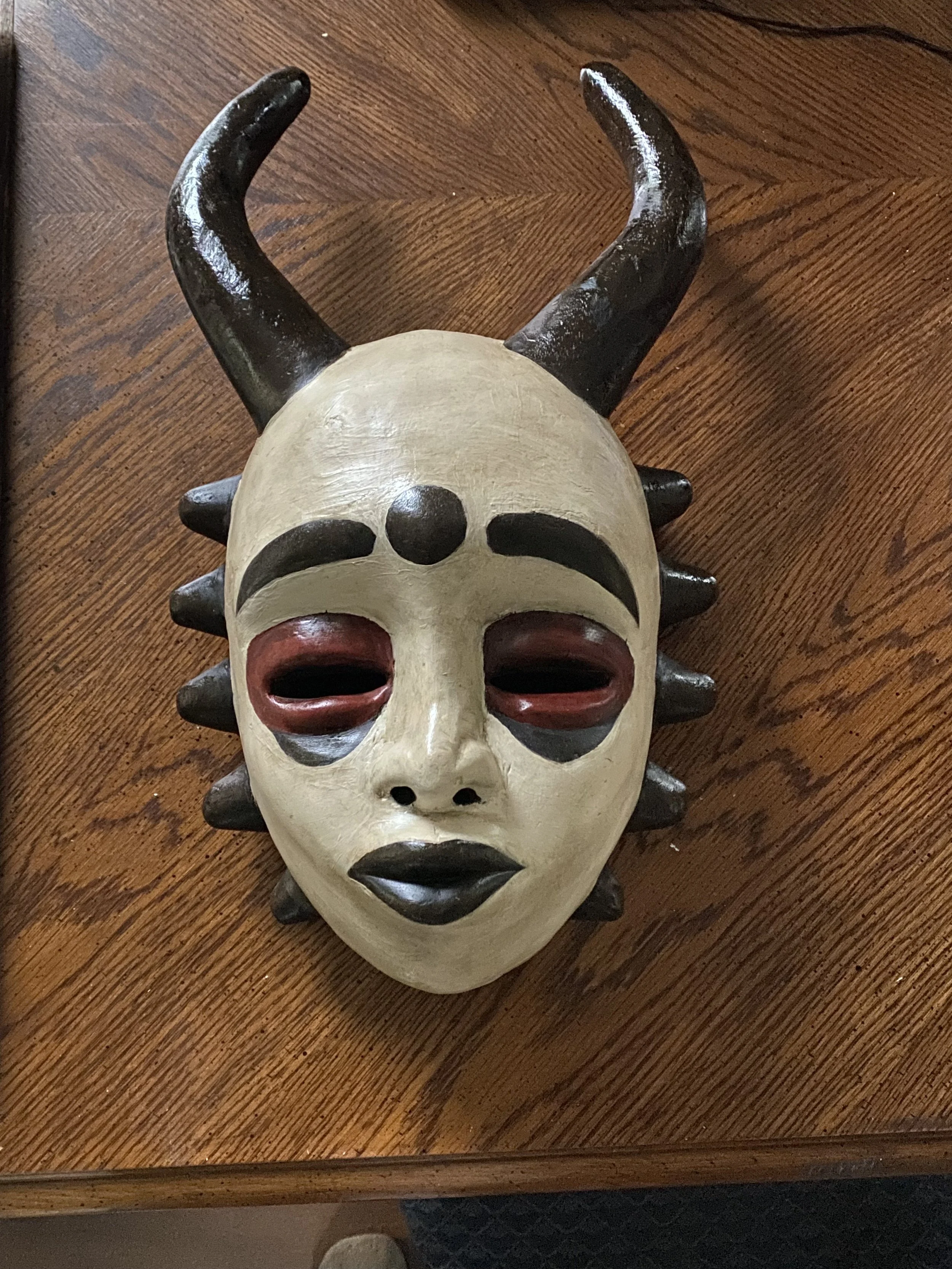 A decorative mask resembling a mythical or tribal face with horns, black lips, and exaggerated facial features, lying on a wooden surface.