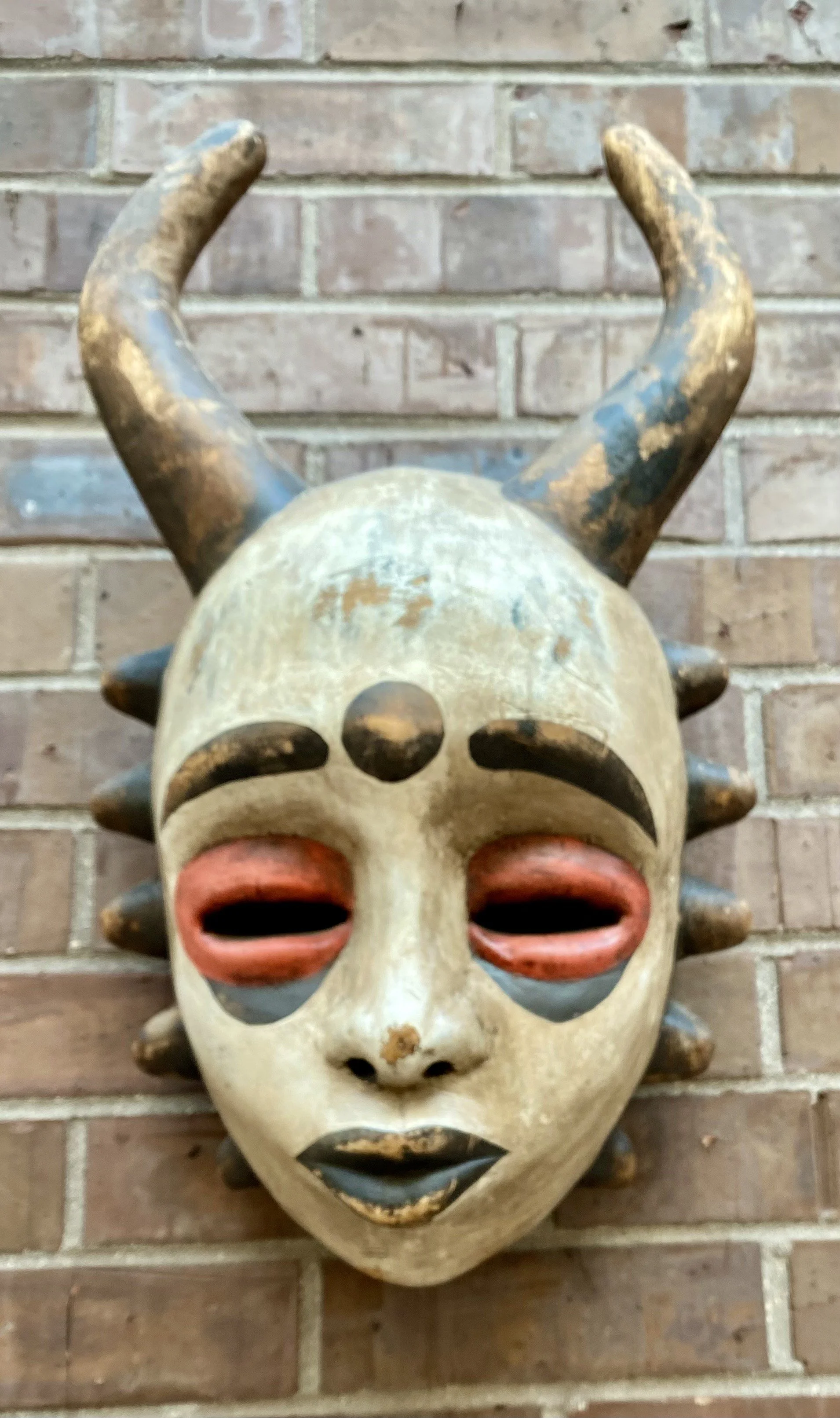 Decorative tribal mask with antler-like horns mounted on a brick wall.