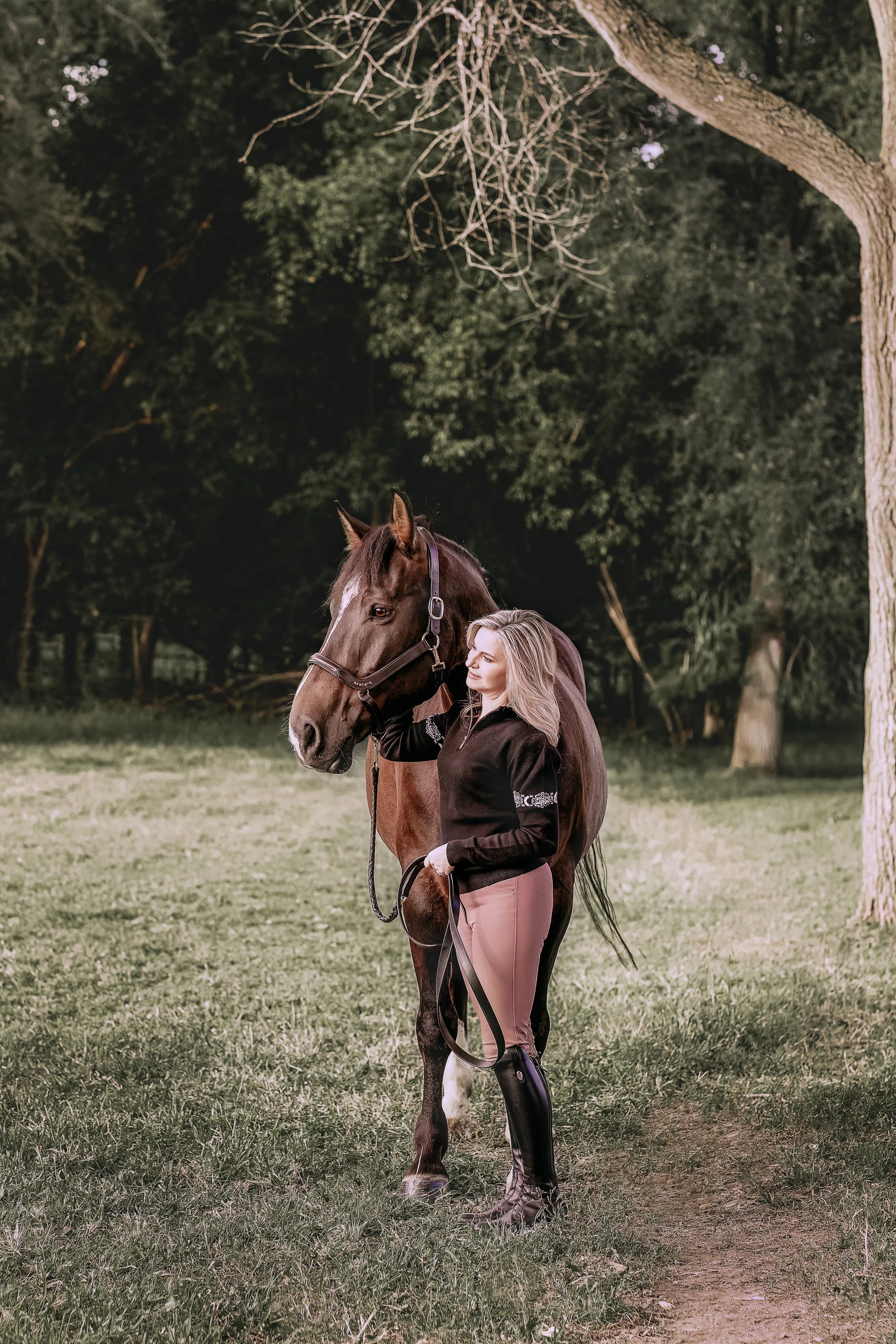 A woman standing next to a brown horse in a grassy area near trees.