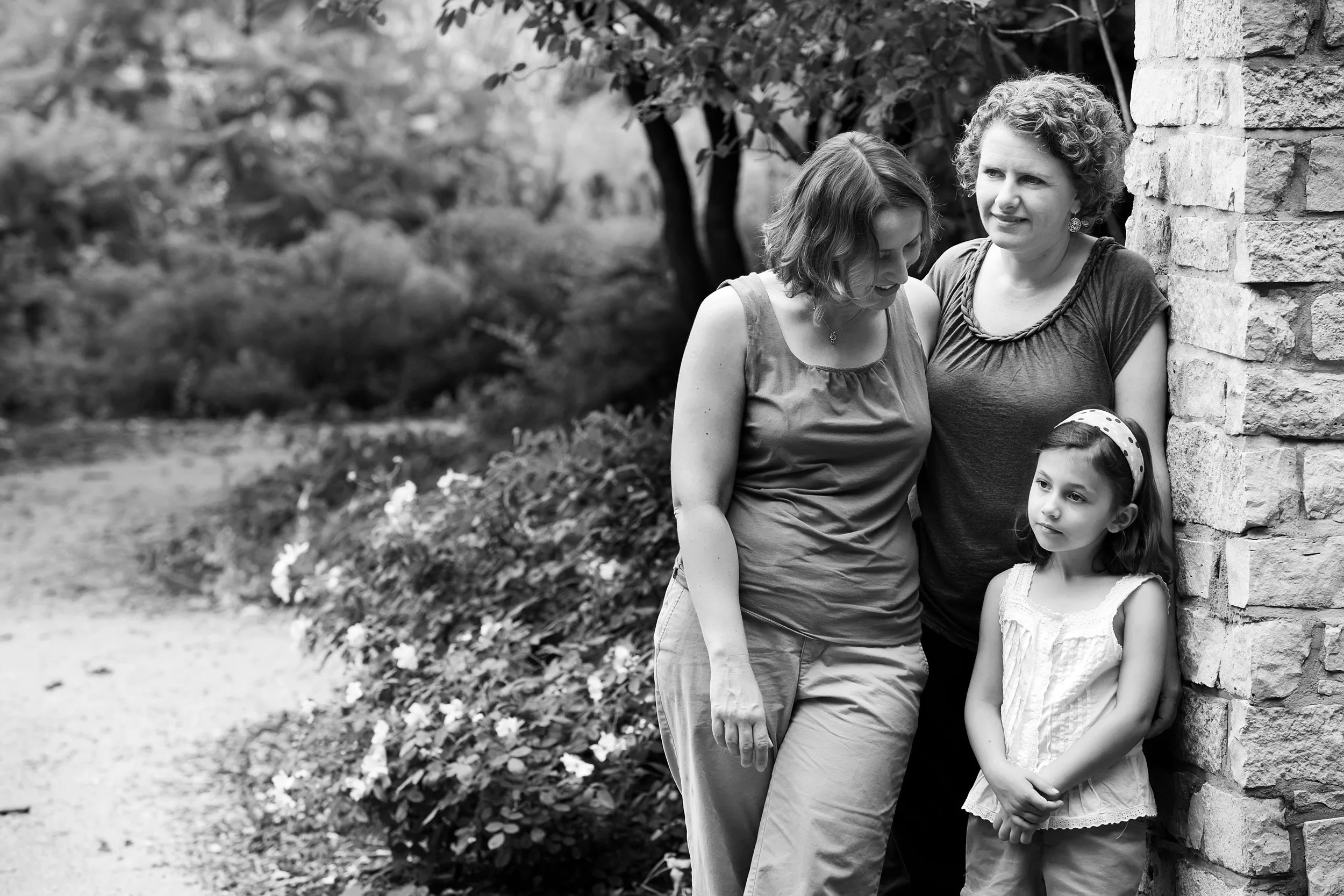 Three females, one adult woman, one teen, and one young girl, standing outdoors beside a stone wall, with a garden and trees in the background.