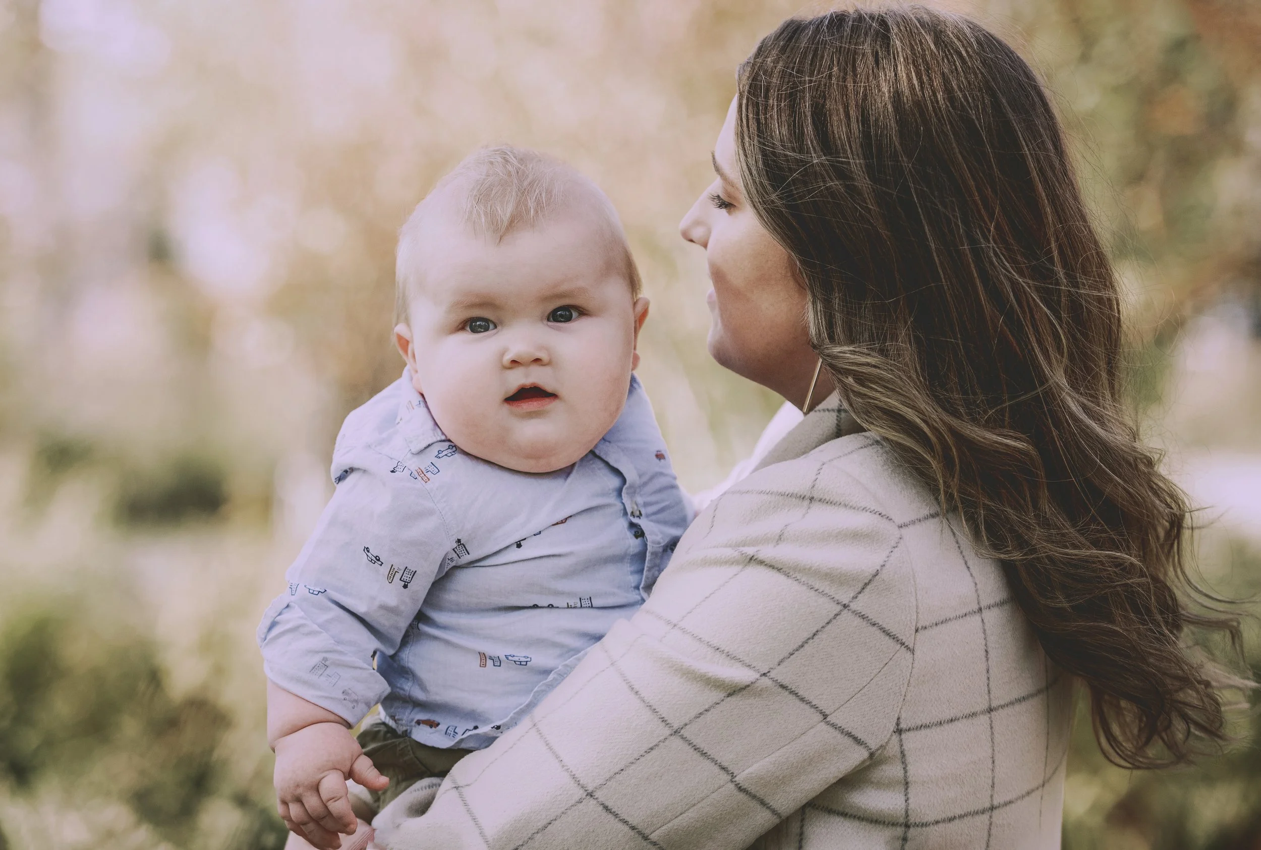 A woman holding a baby outdoors with autumn foliage in the background.