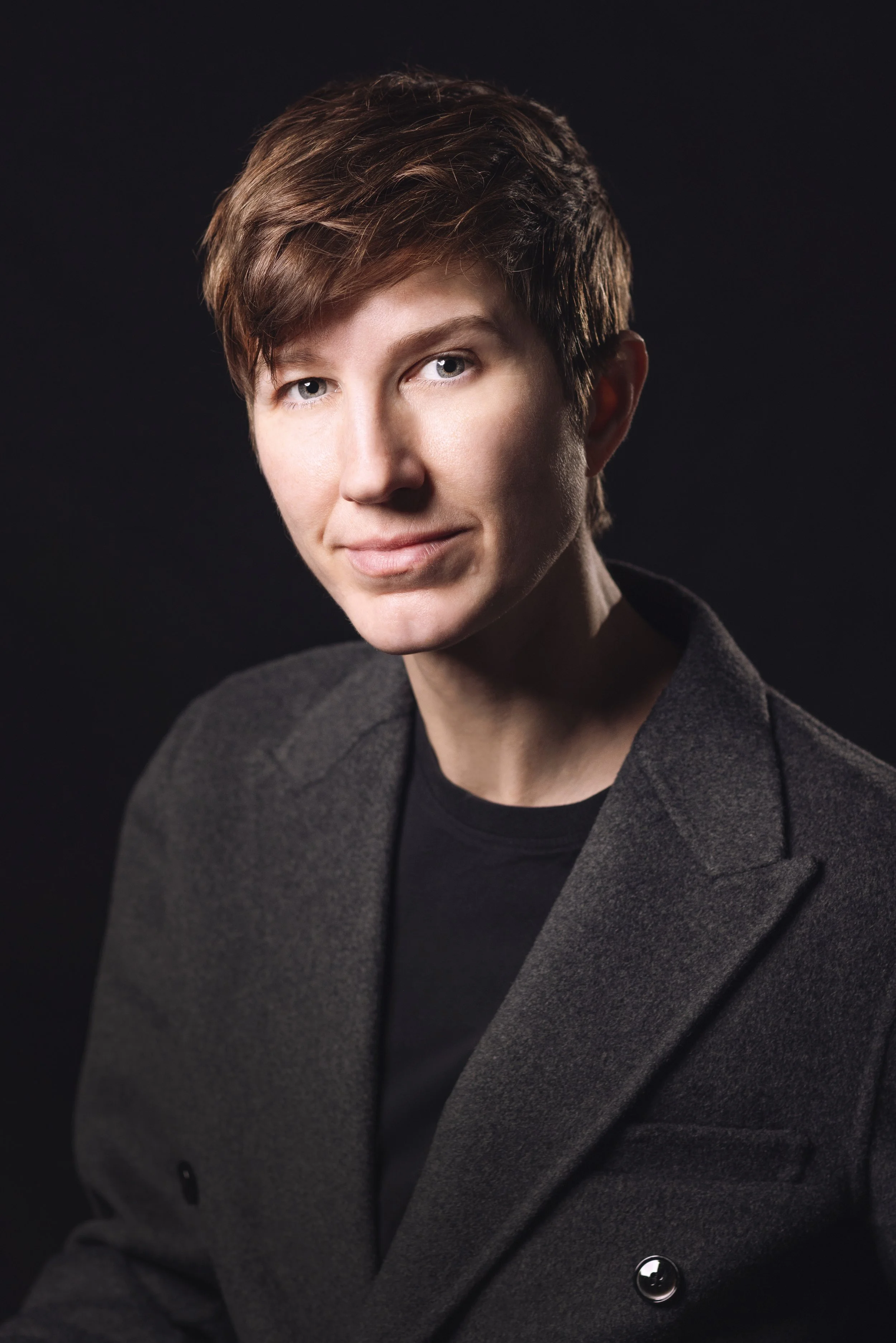 Portrait of a person with short brown hair wearing a dark gray blazer and black shirt, looking at the camera against a black background.