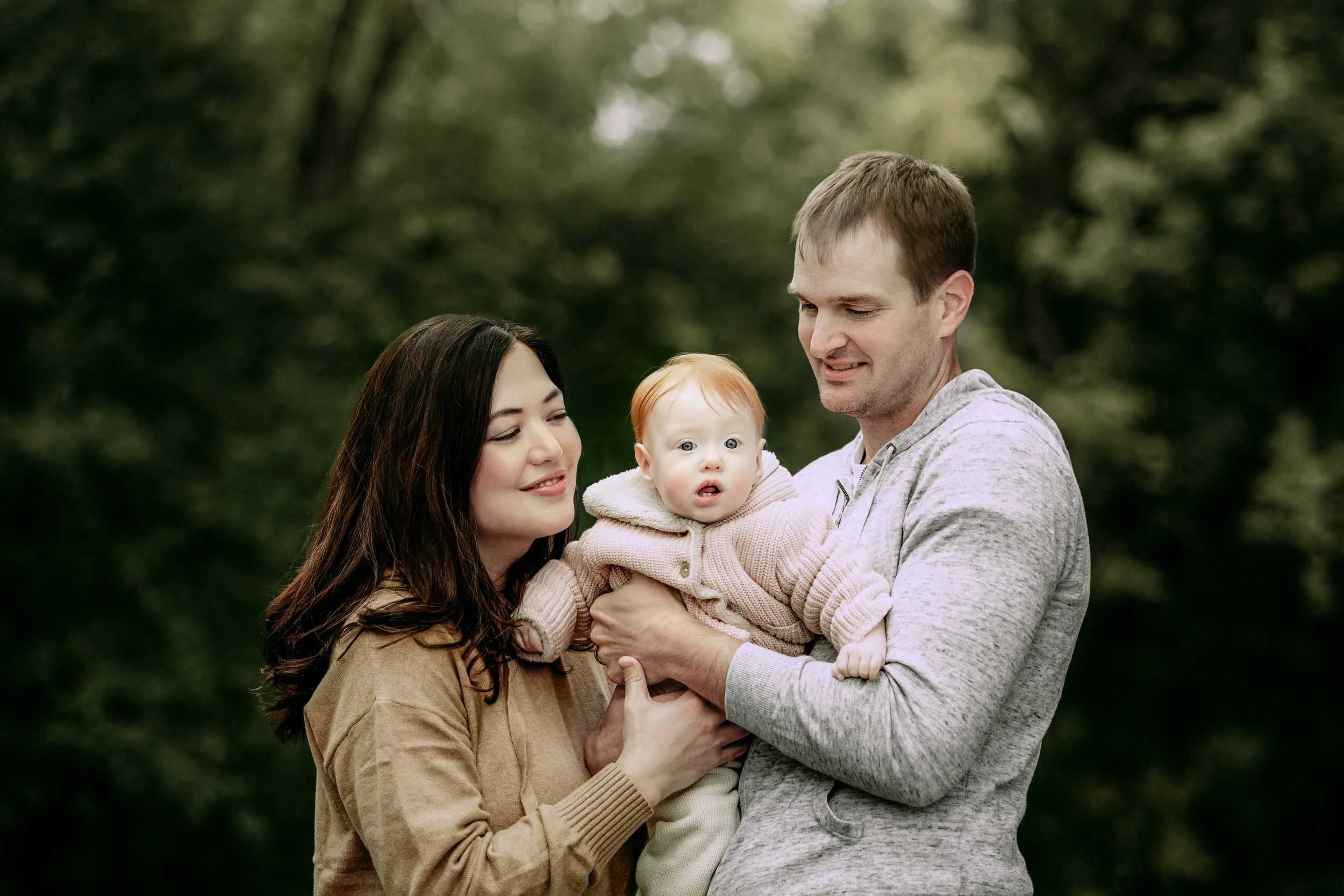 A family of three outdoors, a woman with dark hair, a man, and a baby with red hair, standing in a green, wooded area.