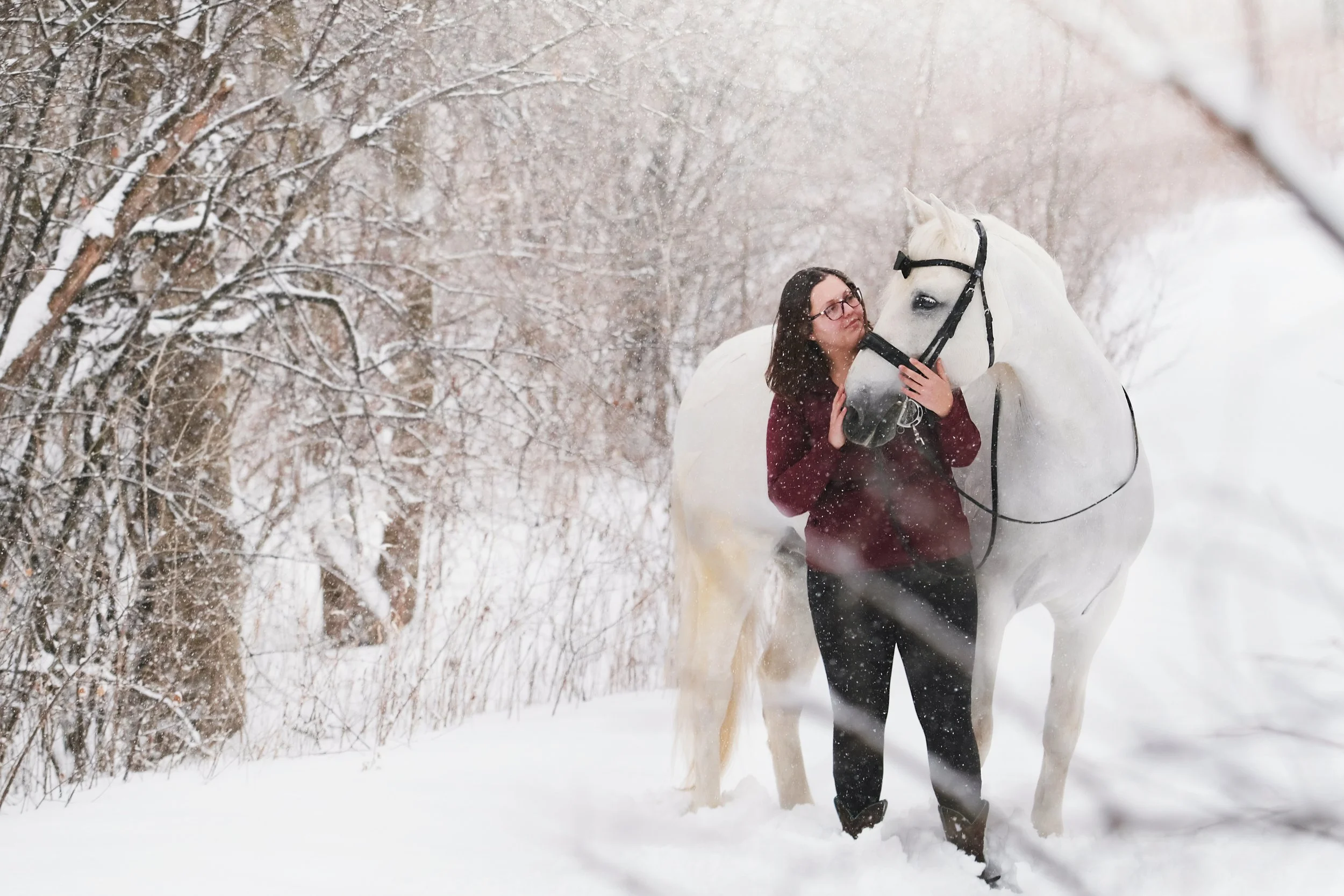 A woman with glasses and a maroon jacket standing in snow, hugging a white horse amidst falling snow, with snow-covered trees in the background.