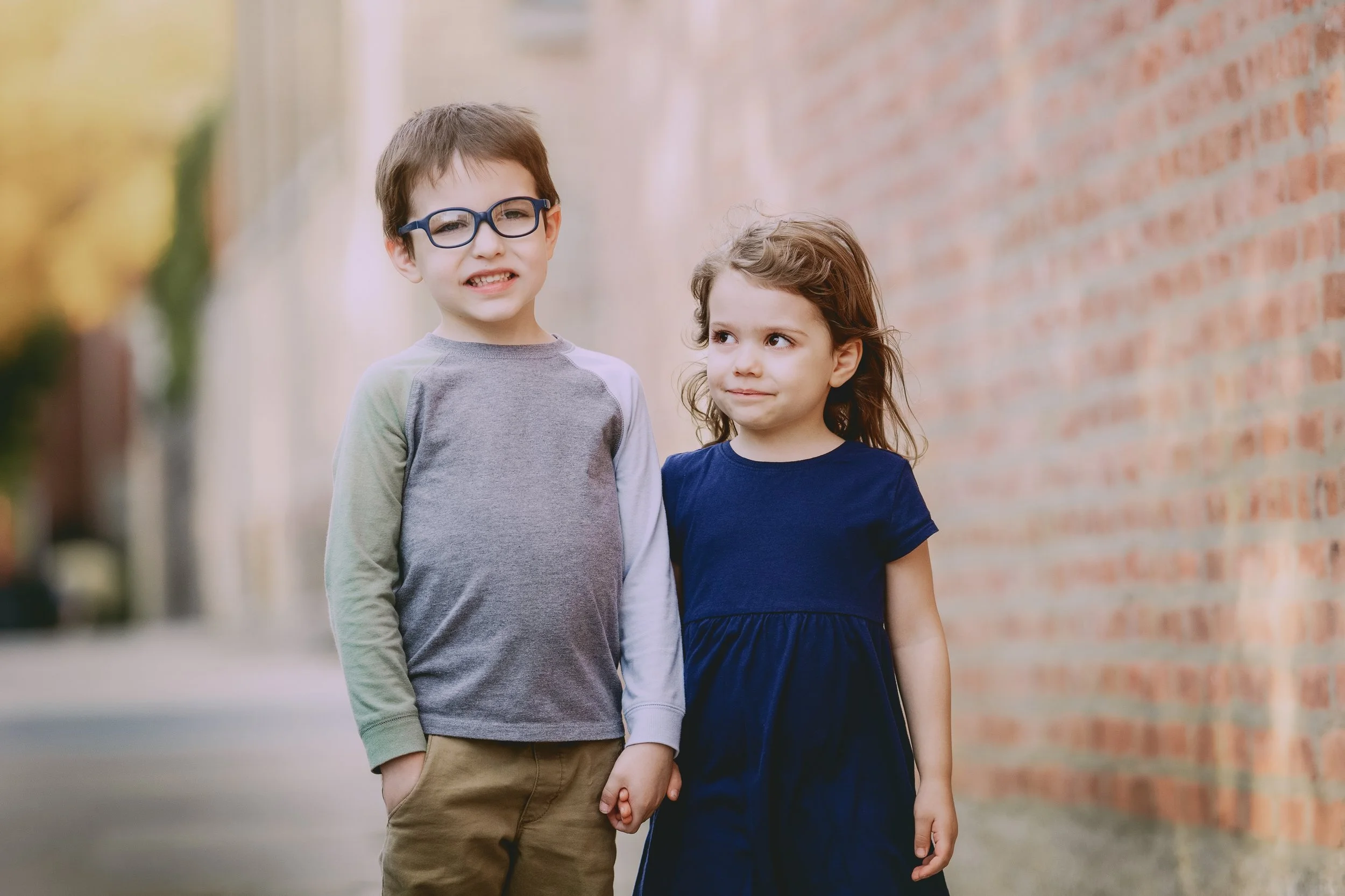 A young boy and girl holding hands outdoors, standing on a sidewalk near a brick wall, with blurred trees in the background.