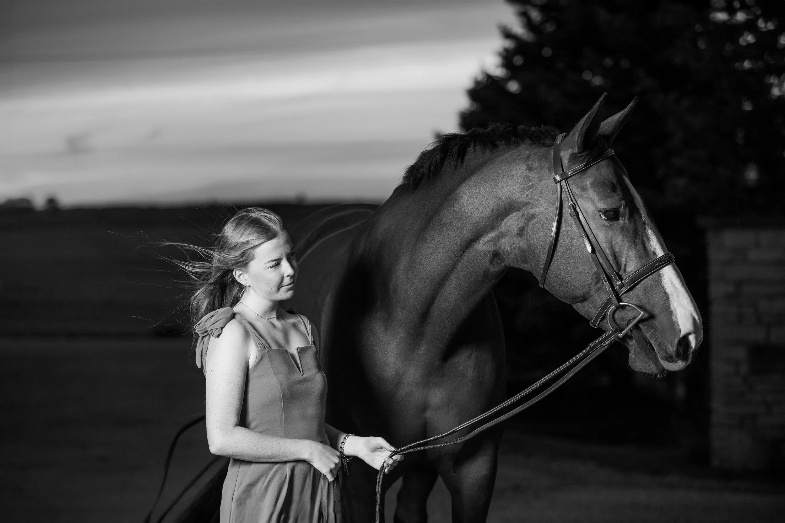 Black and white photo of a young woman with long hair walking a horse outdoors during sunset or dusk.