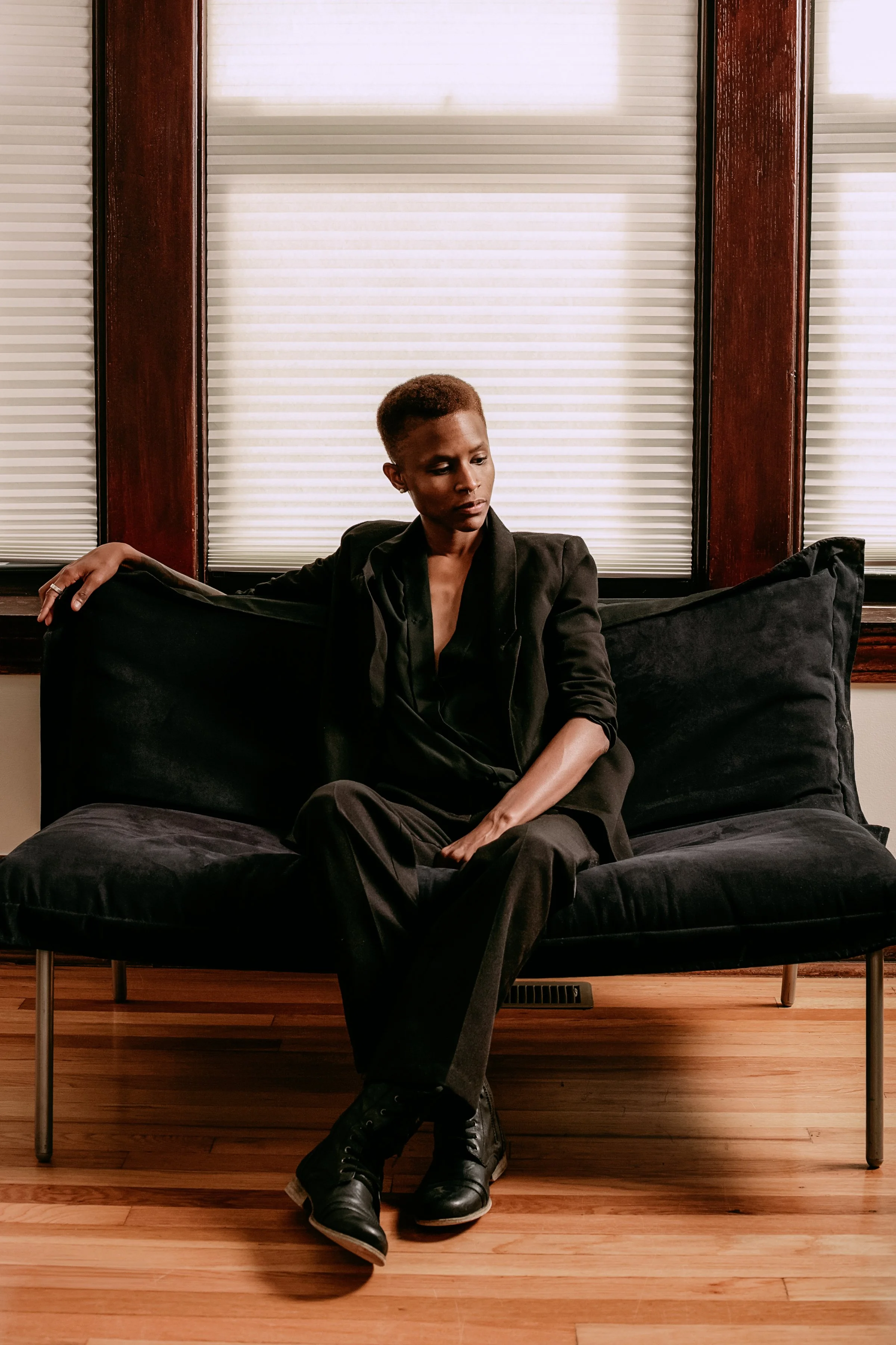 A woman with short hair dressed in black, sitting on a black sofa in a room with hardwood floors and windows with closed blinds.