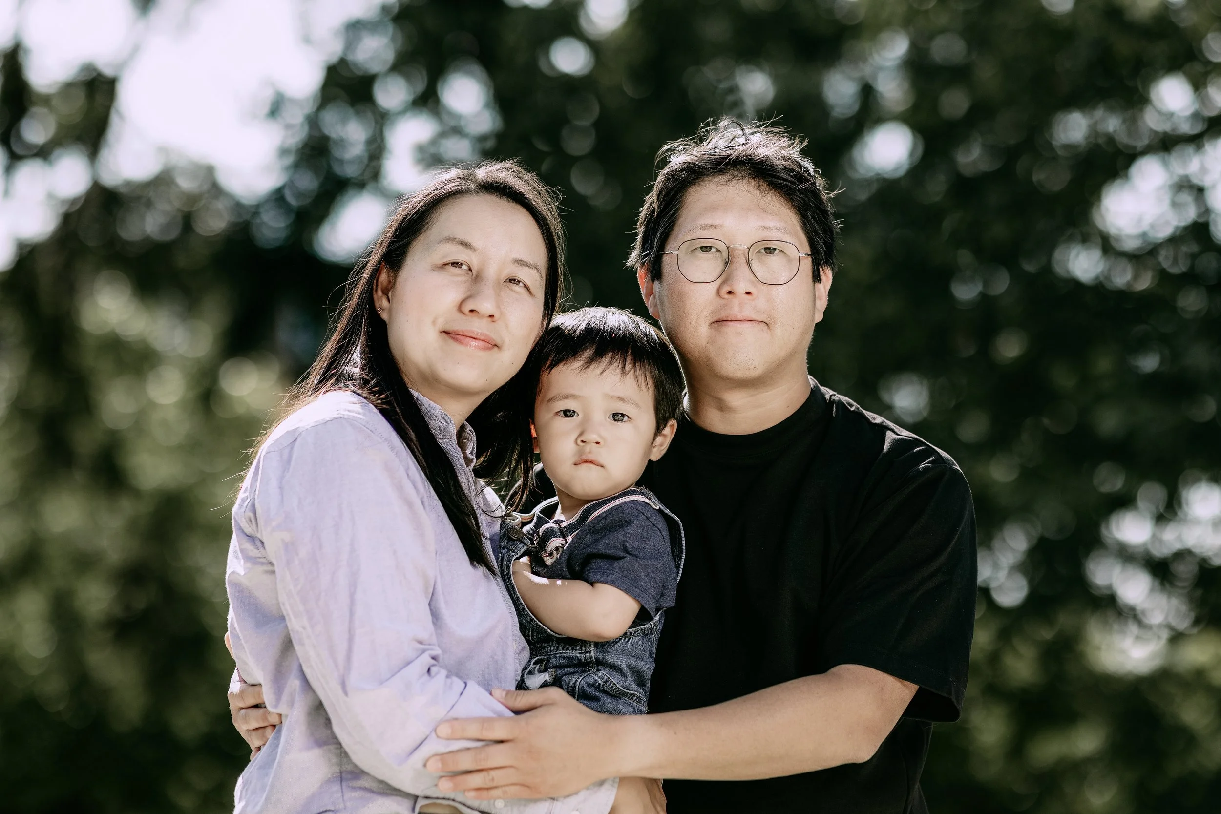 A family of three standing outdoors, with trees in the background, smiling and embracing each other.