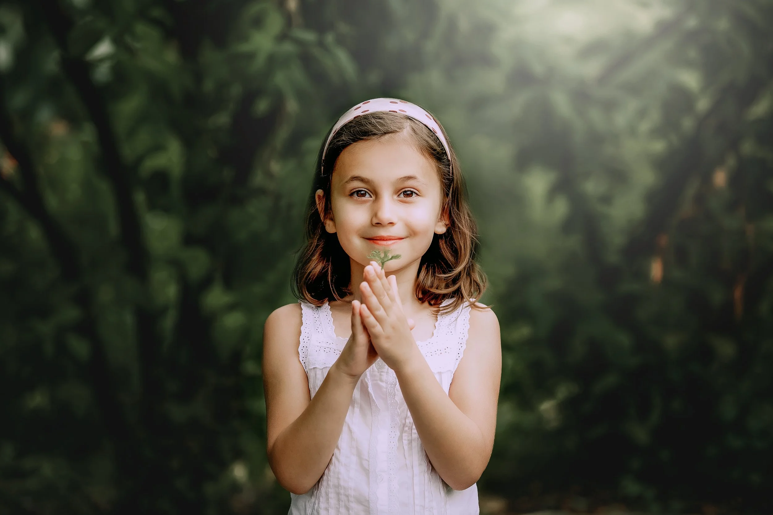 A young girl with brown hair, wearing a white dress and pink headband, holds a small green leaf with her hands clasped in front of her chest, standing outdoors in a lush green forest.