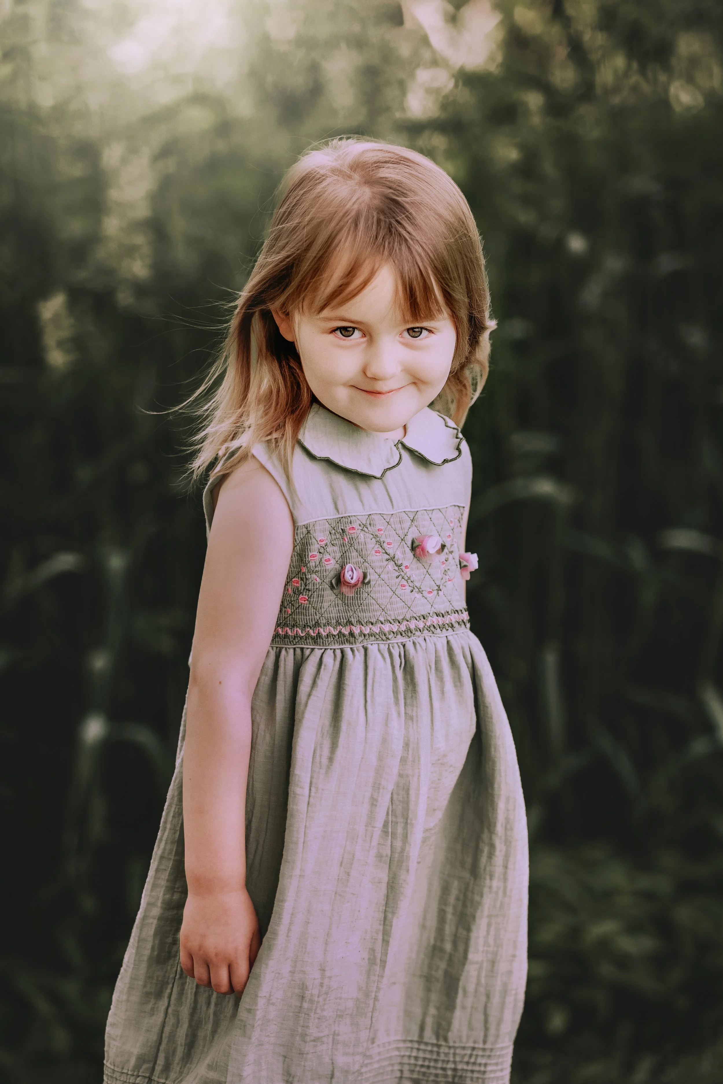 A young girl with light brown hair and fair skin standing outdoors in a field of green plants, smiling softly at the camera.