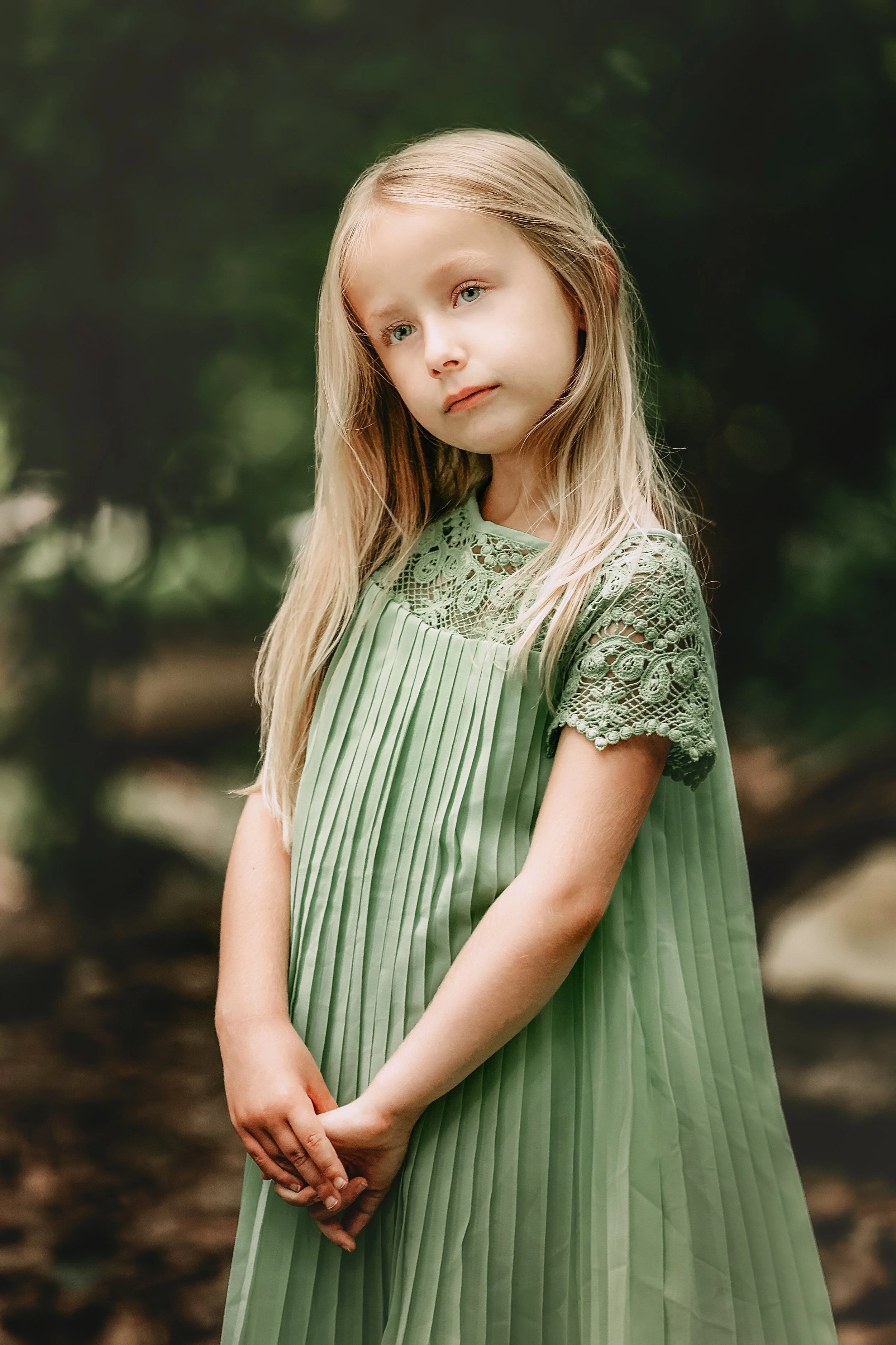 A young girl with long blonde hair wearing a green pleated dress with lace details on the top and sleeves, standing outdoors with a blurred green natural background.
