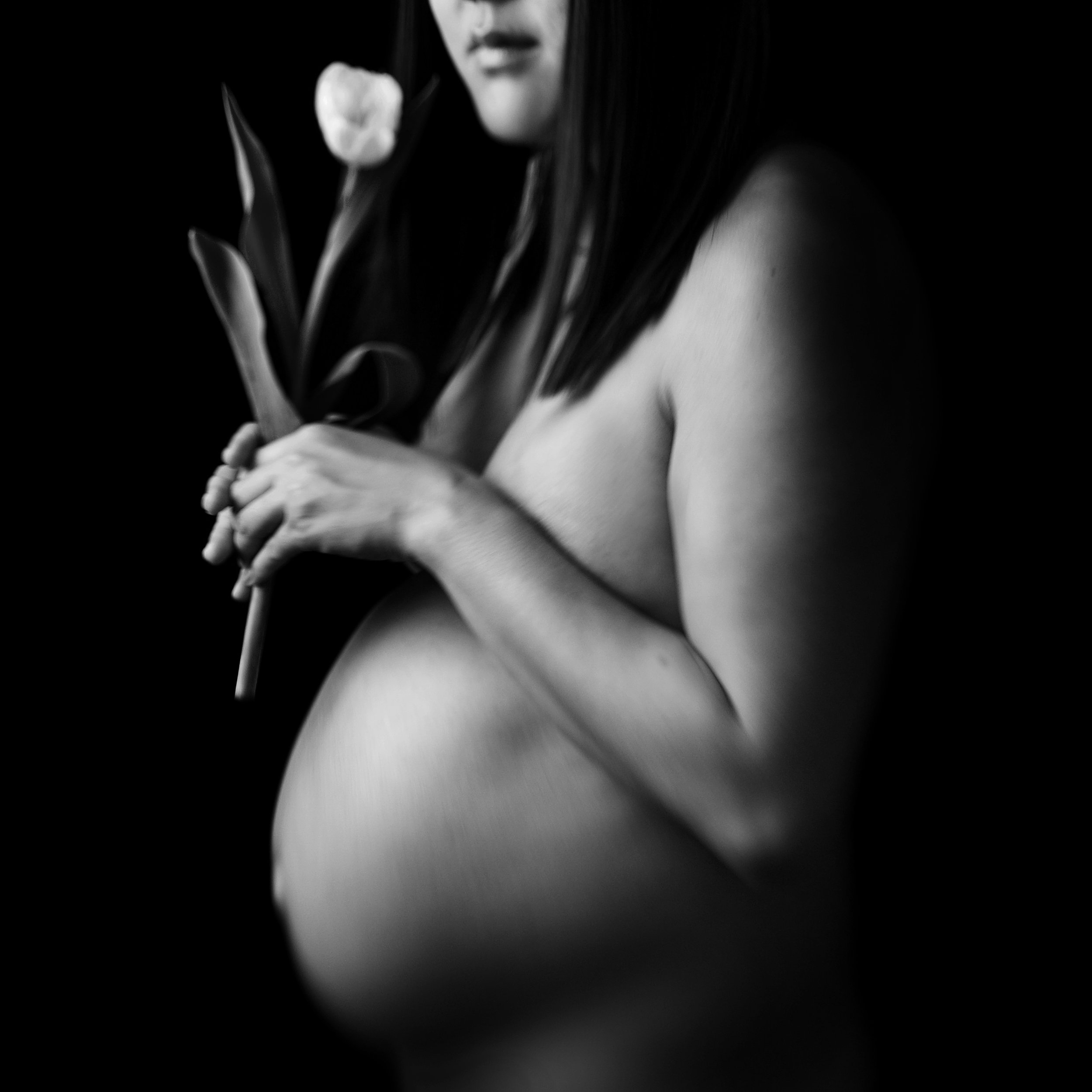 A black and white photo of a pregnant woman holding a flower close to her face.