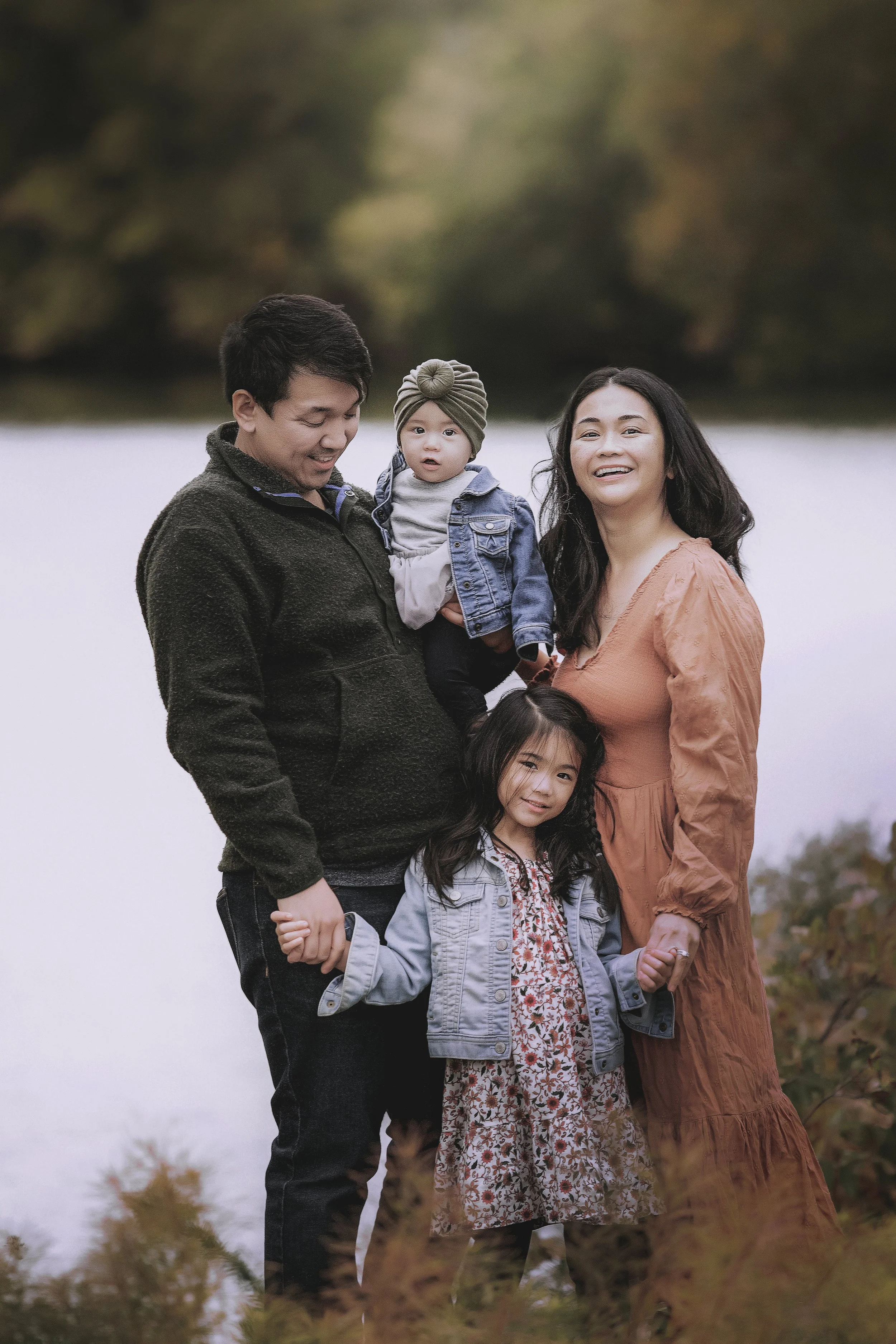 A family of four standing outdoors near a river or lake during autumn, holding hands and smiling. The father is holding a toddler, and the mother is standing beside a young girl, with trees in the background.