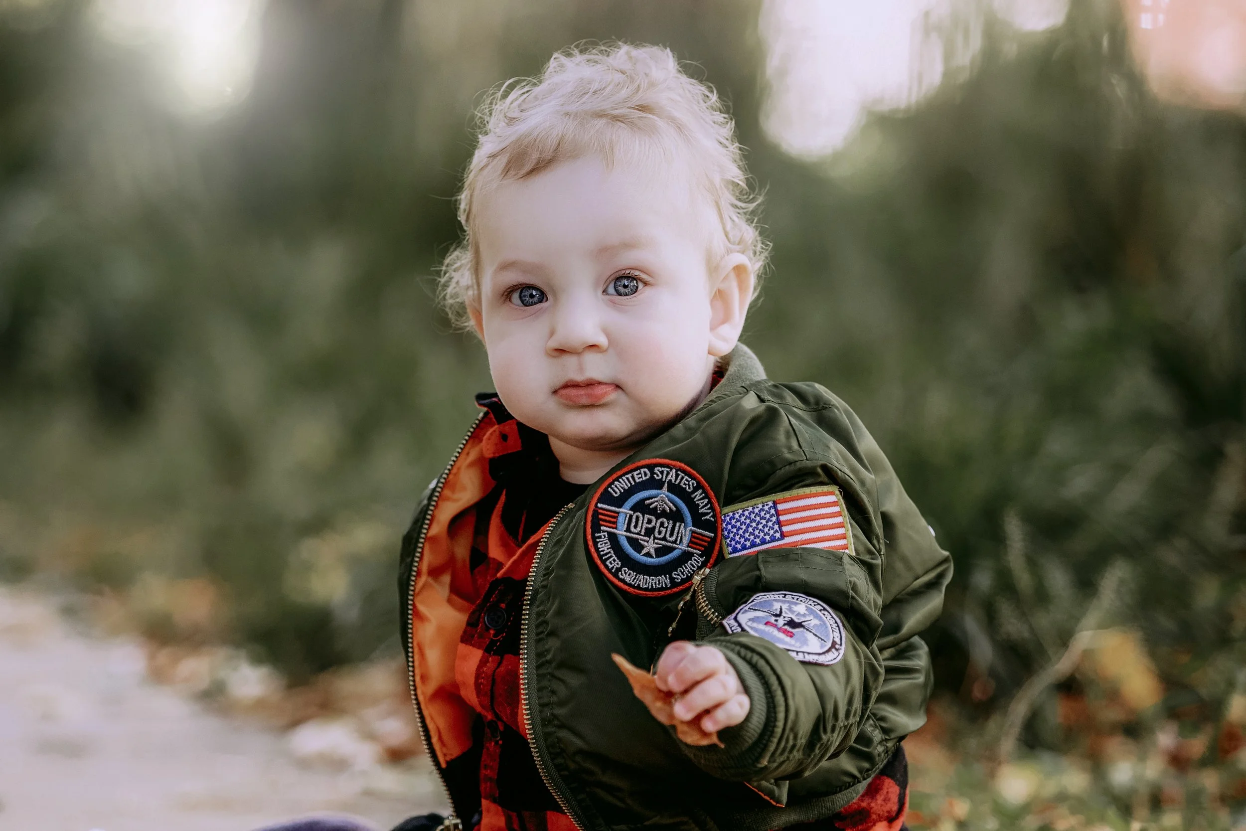 A young child with blonde hair and blue eyes wearing a green military-style bomber jacket with patches, holding a small object, outdoors with trees and blurred background.