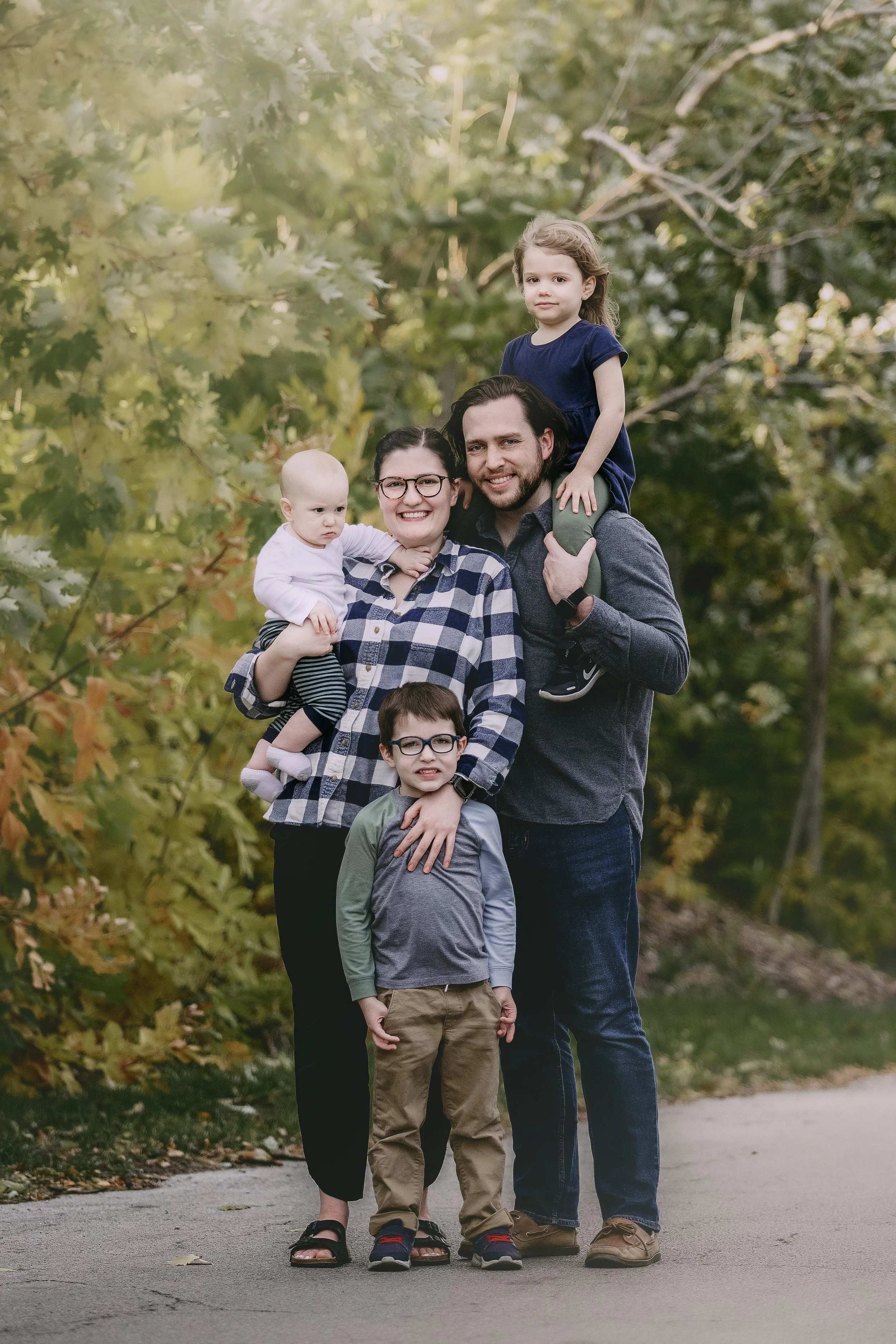Family of five outdoors, two children and two adults smiling, standing on a trail surrounded by fall foliage.