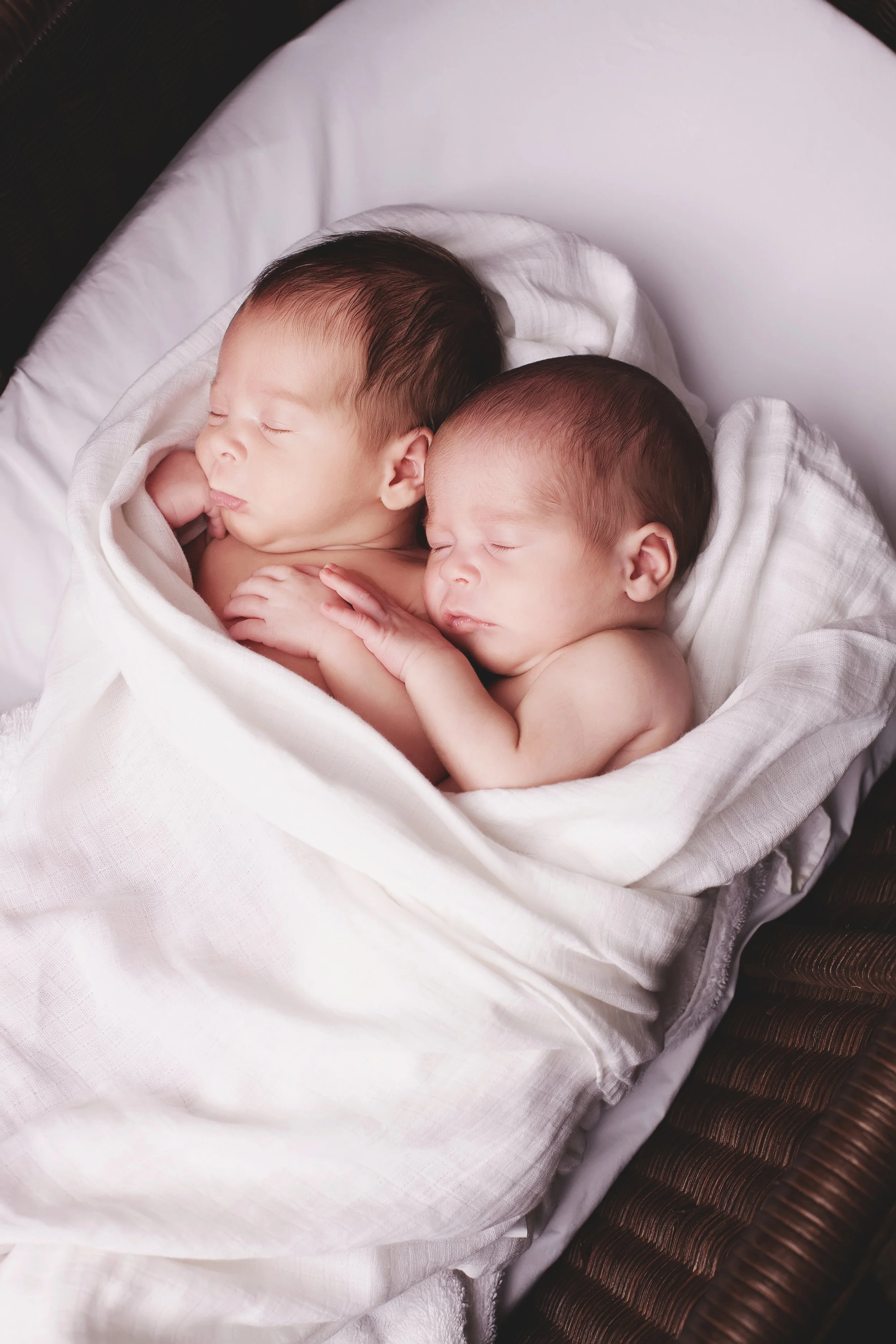 Two sleeping newborn babies wrapped in a white blanket, lying close together in a bassinet.