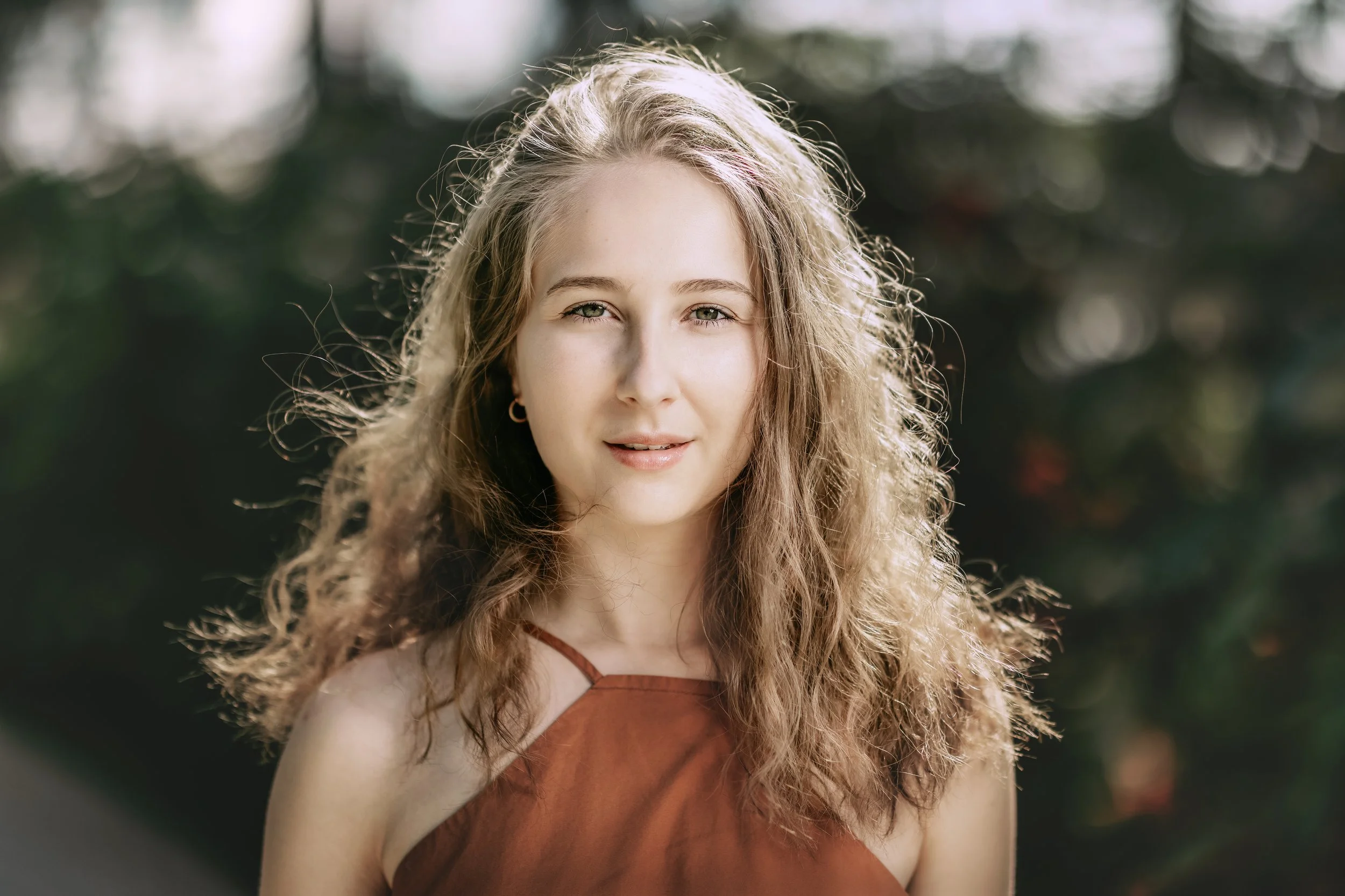 A woman with curly blonde hair, wearing a rust-colored top, outdoors with a blurred green background.