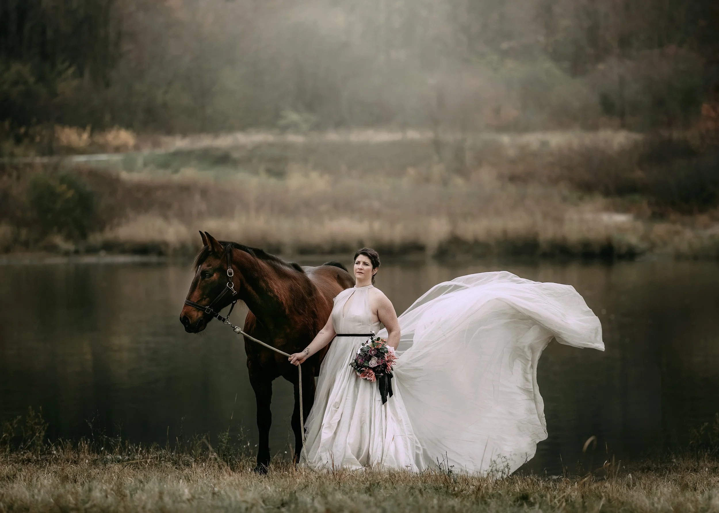 A woman in a white wedding dress standing next to a brown horse by a lake, holding a bouquet of flowers, with a scenic background of trees and water.