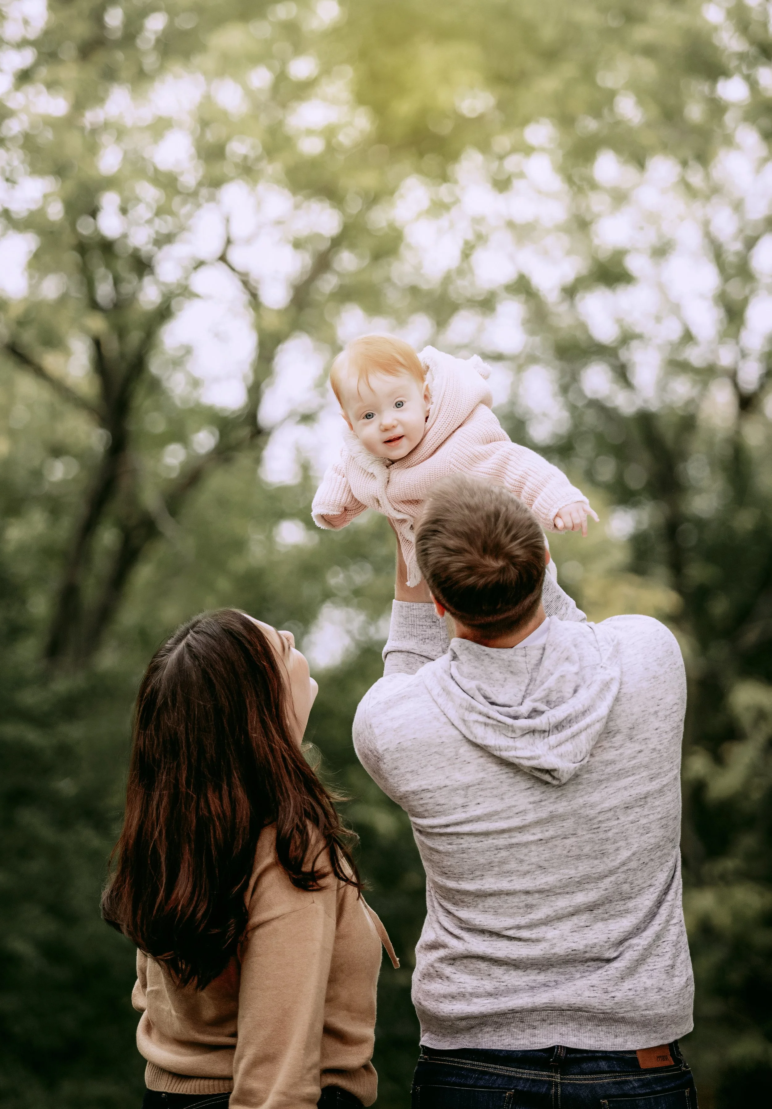 A man lifting a baby girl into the air outdoors, with a woman smiling nearby, surrounded by green trees.