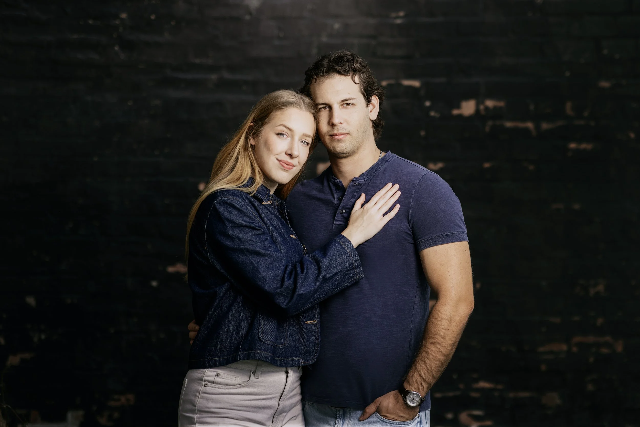 A young couple standing close together against a dark background, with the woman resting her hand on the man's chest.