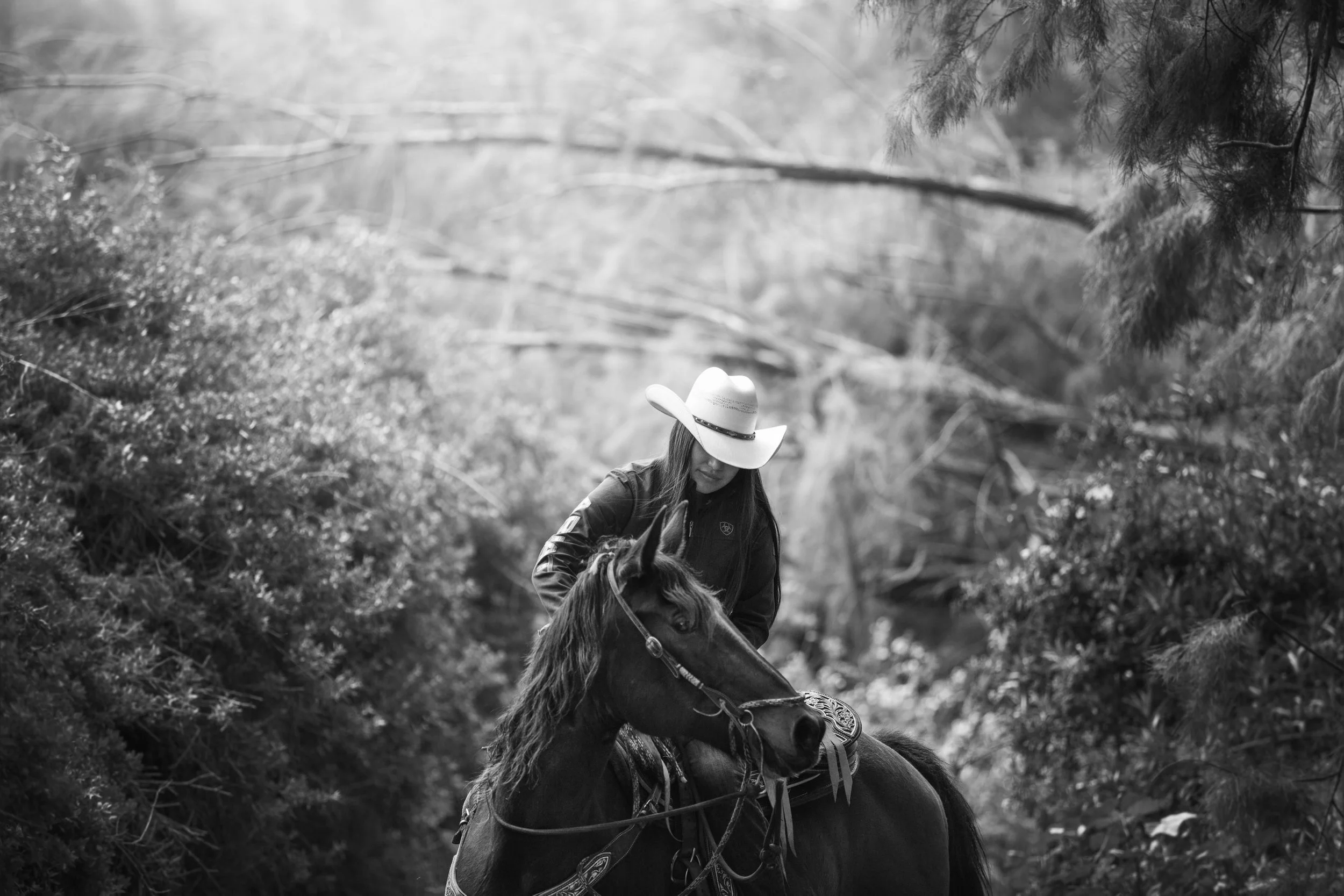 A woman wearing a cowboy hat riding a horse, surrounded by trees and bushes, in black and white.