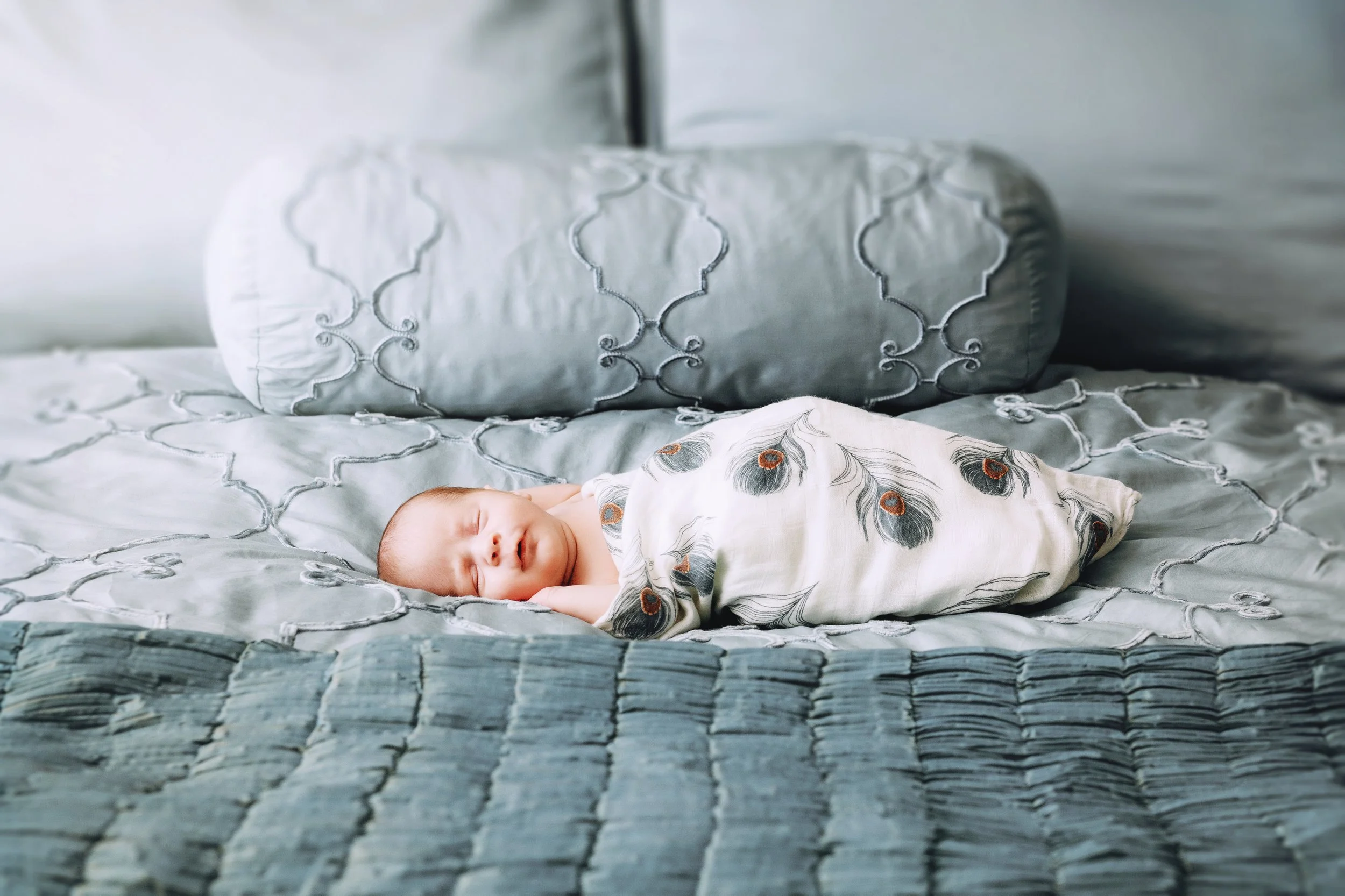 A sleeping baby wrapped in a white blanket with peacock feather patterns, lying on a bed with gray embroidered bedding and pillows.