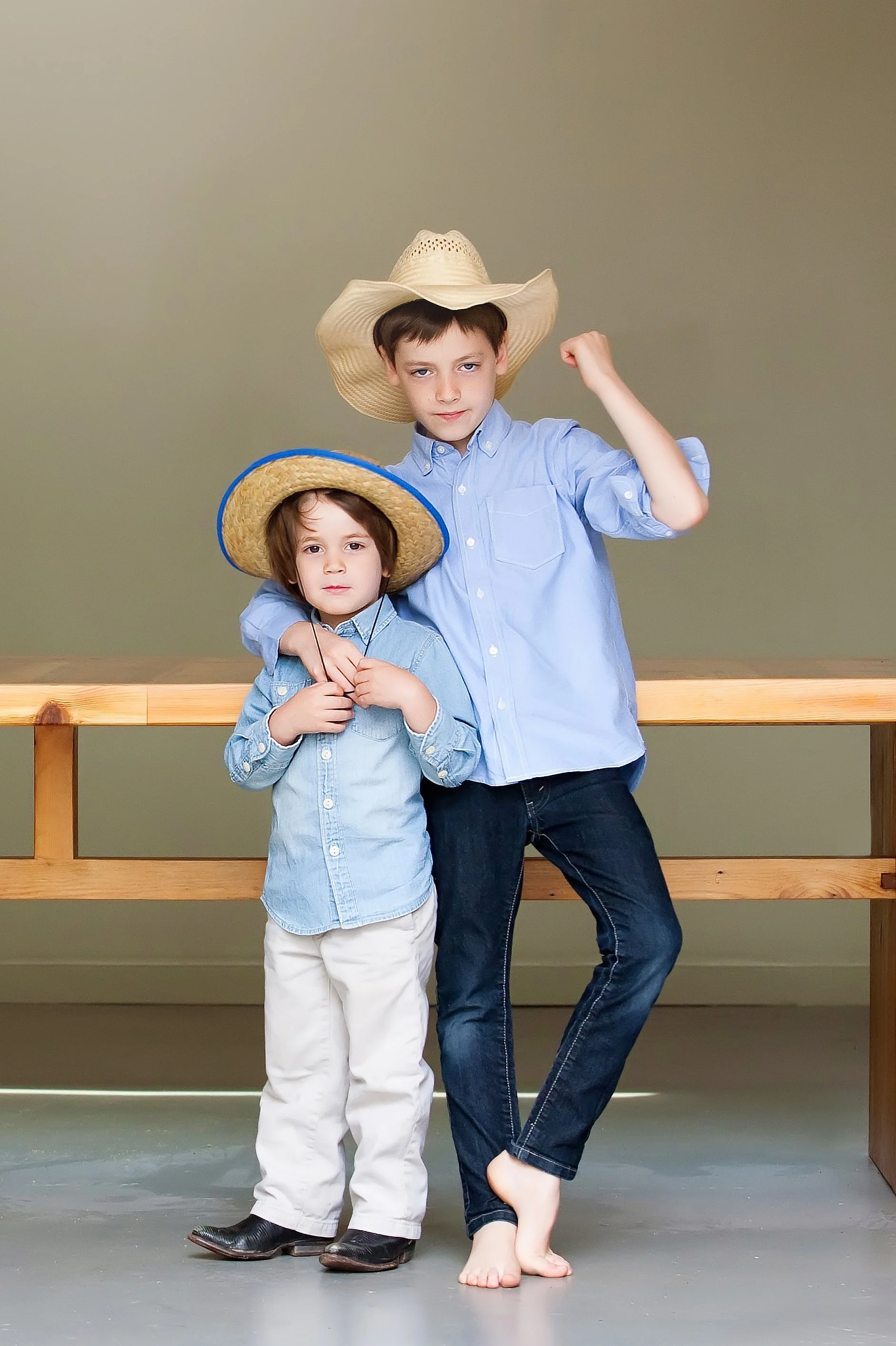 Two boys wearing cowboy hats and denim shirts, standing by a wooden rail. One boy is barefoot, flexing his arm, and the other boy is wearing cowboy boots.