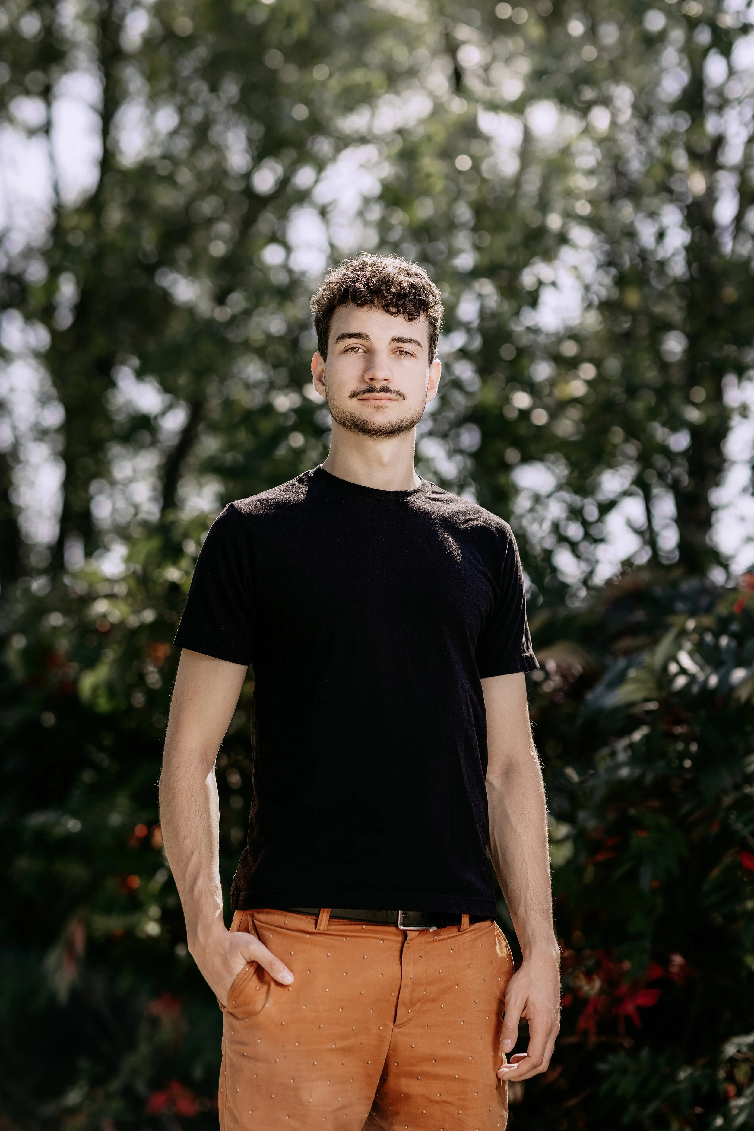 Young man with curly hair, wearing a black t-shirt and orange pants, standing outdoors with a background of trees and foliage.