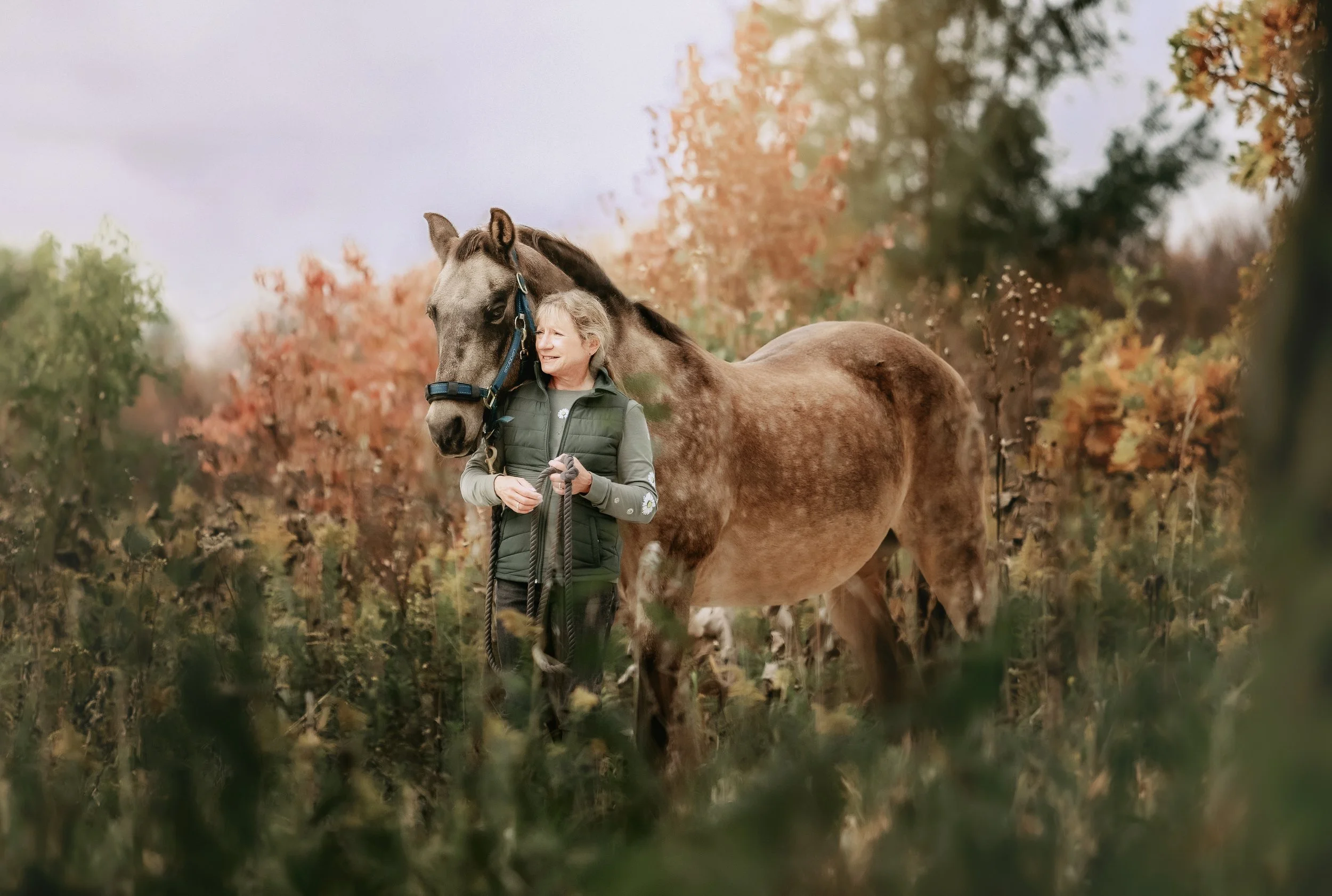 An older woman in a green vest standing in an autumn-colored forest, smiling while holding a leash attached to a brown horse with a blue halter, both sharing a moment of companionship.
