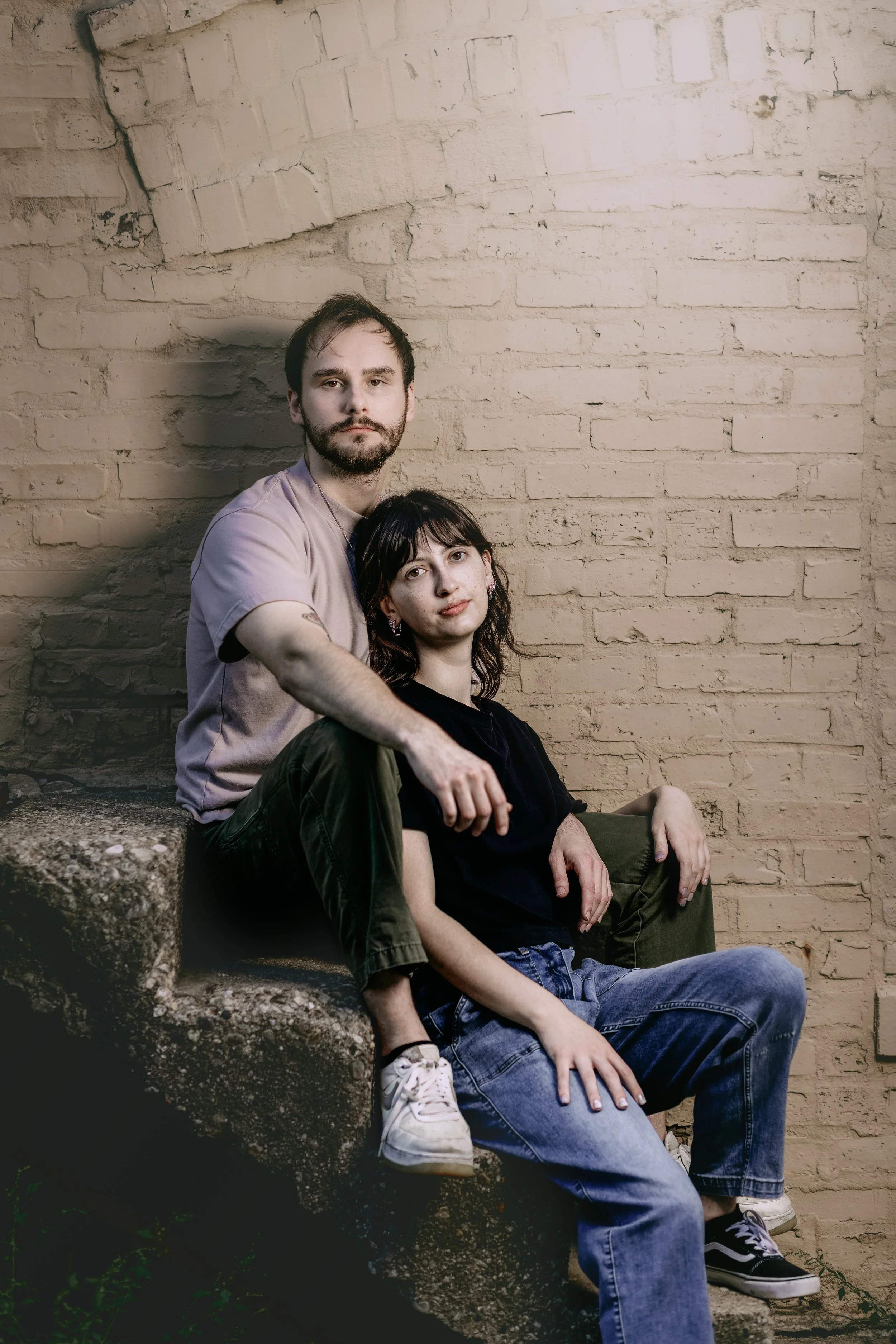 A man and a woman sitting close together against a beige brick wall. The man has a beard and mustache, and the woman has dark hair and is wearing a black top and jeans. The man is seated on a stone staircase with the woman sitting on his lap, and bot