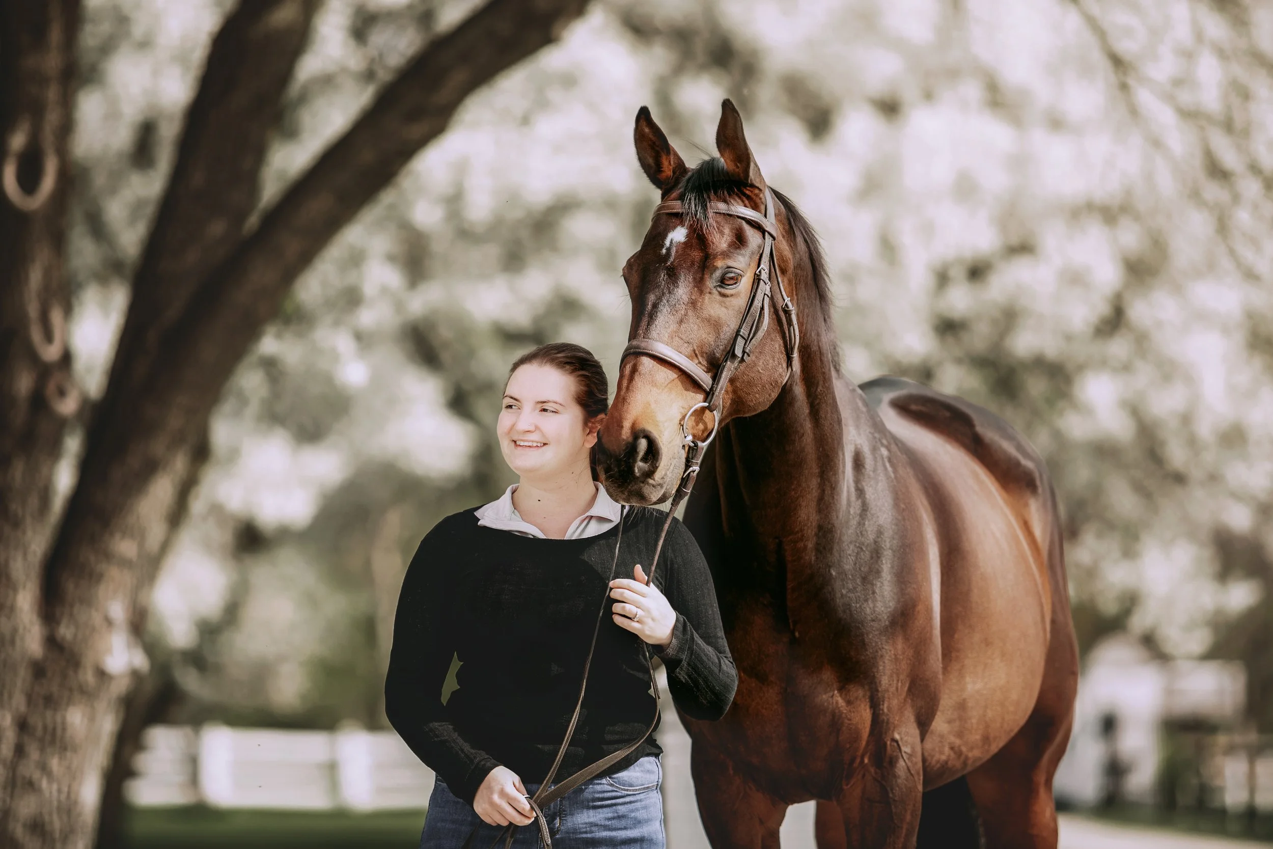 A young woman smiling and standing next to a brown horse under a large tree with blurred background.